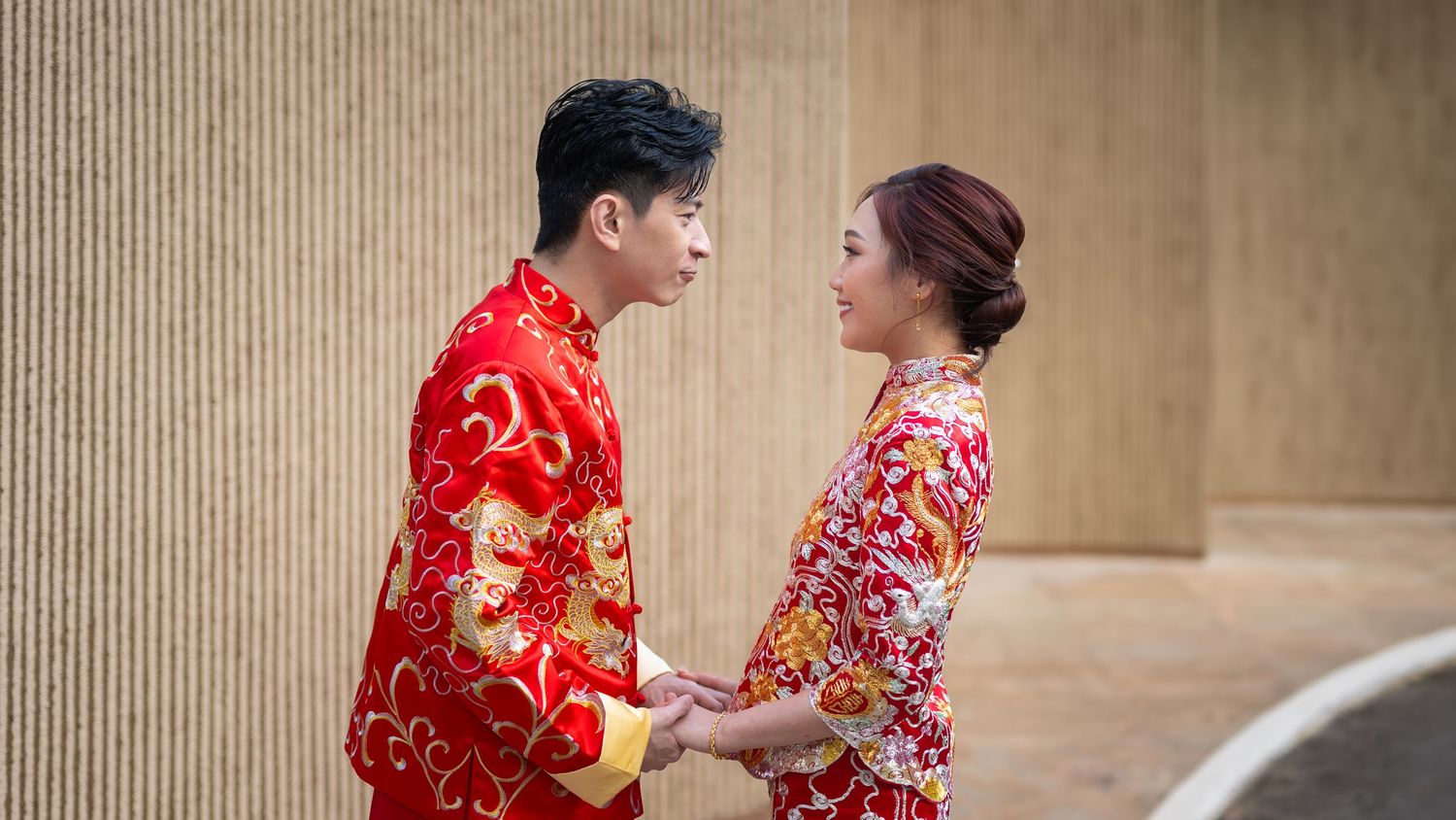 A couple in traditional Chinese red wedding attire holds hands and gazes at each other outdoors.