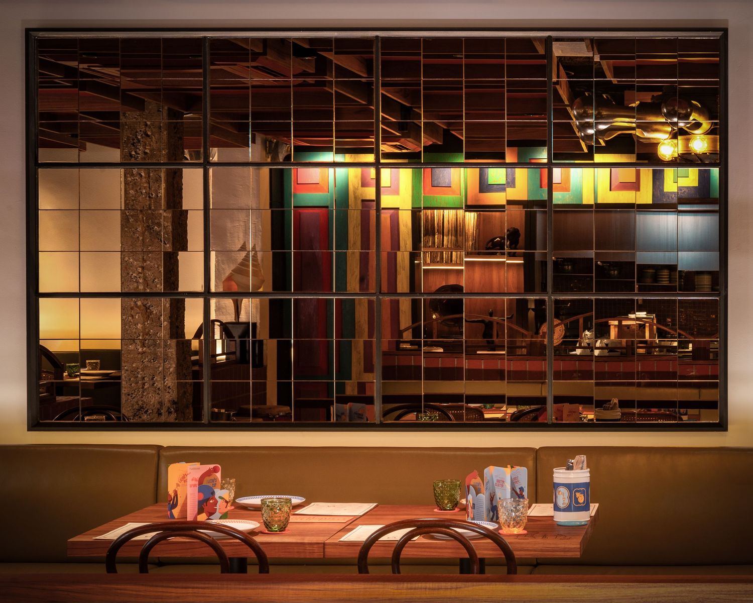Warm restaurant interior viewed through large grid window with wooden dining table and colorful artwork on walls.