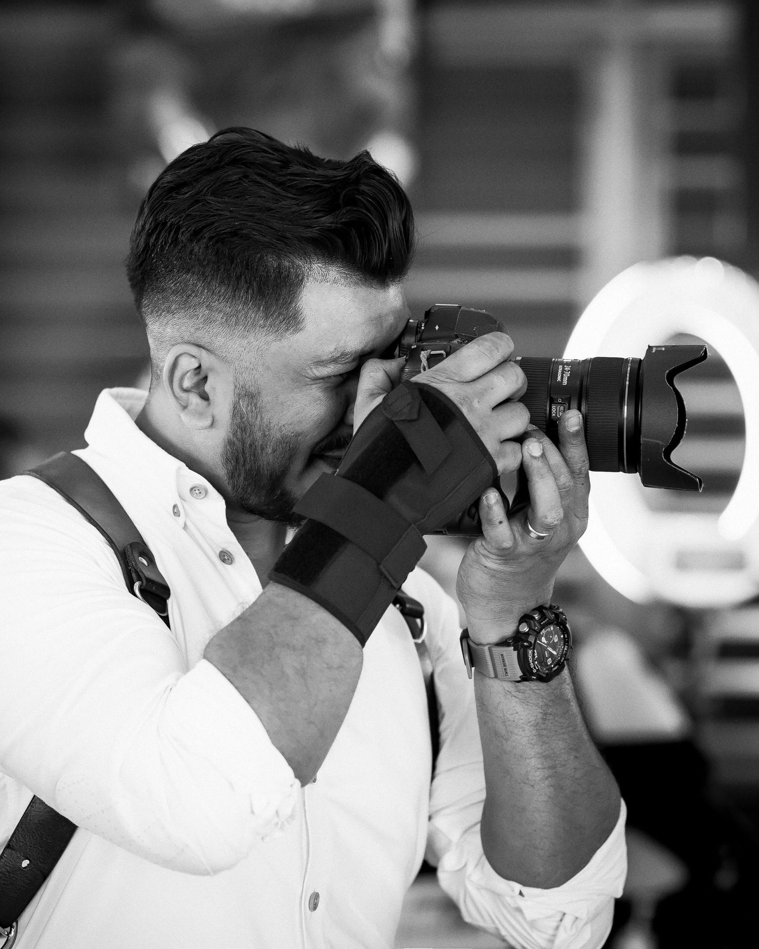 Bearded male photographer in white shirt capturing photos with professional DSLR camera in black and white.