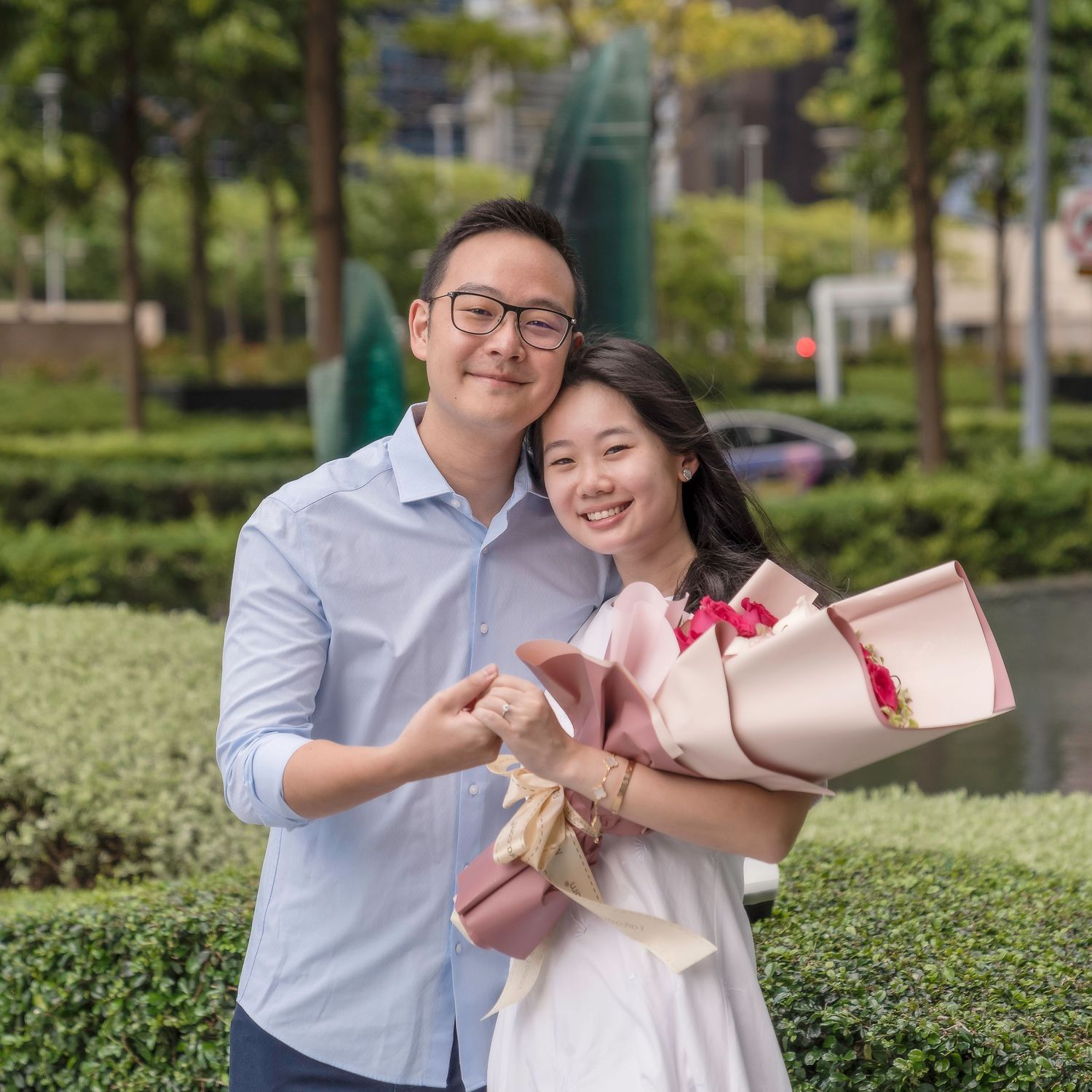 Couple smiling outdoors, man hugging woman holding pink flower bouquet, green hedges background.