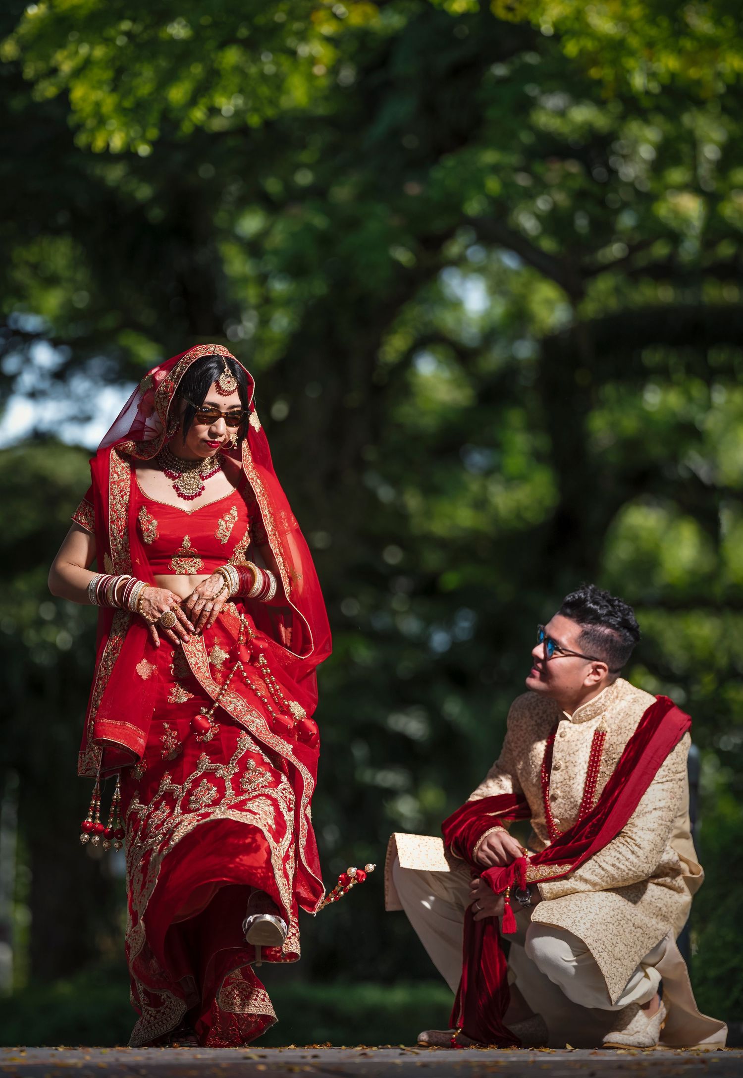 Indian bride in red lehenga and groom in cream sherwani share a romantic moment outdoors during traditional wedding.