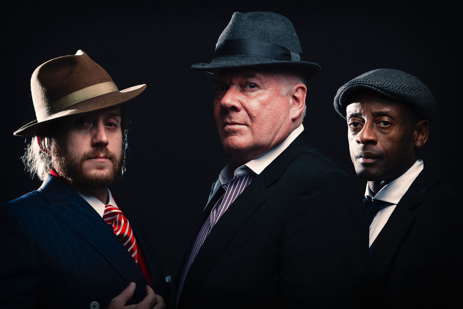 Three men in stylish fedora hats and suits pose dramatically against a dark background.