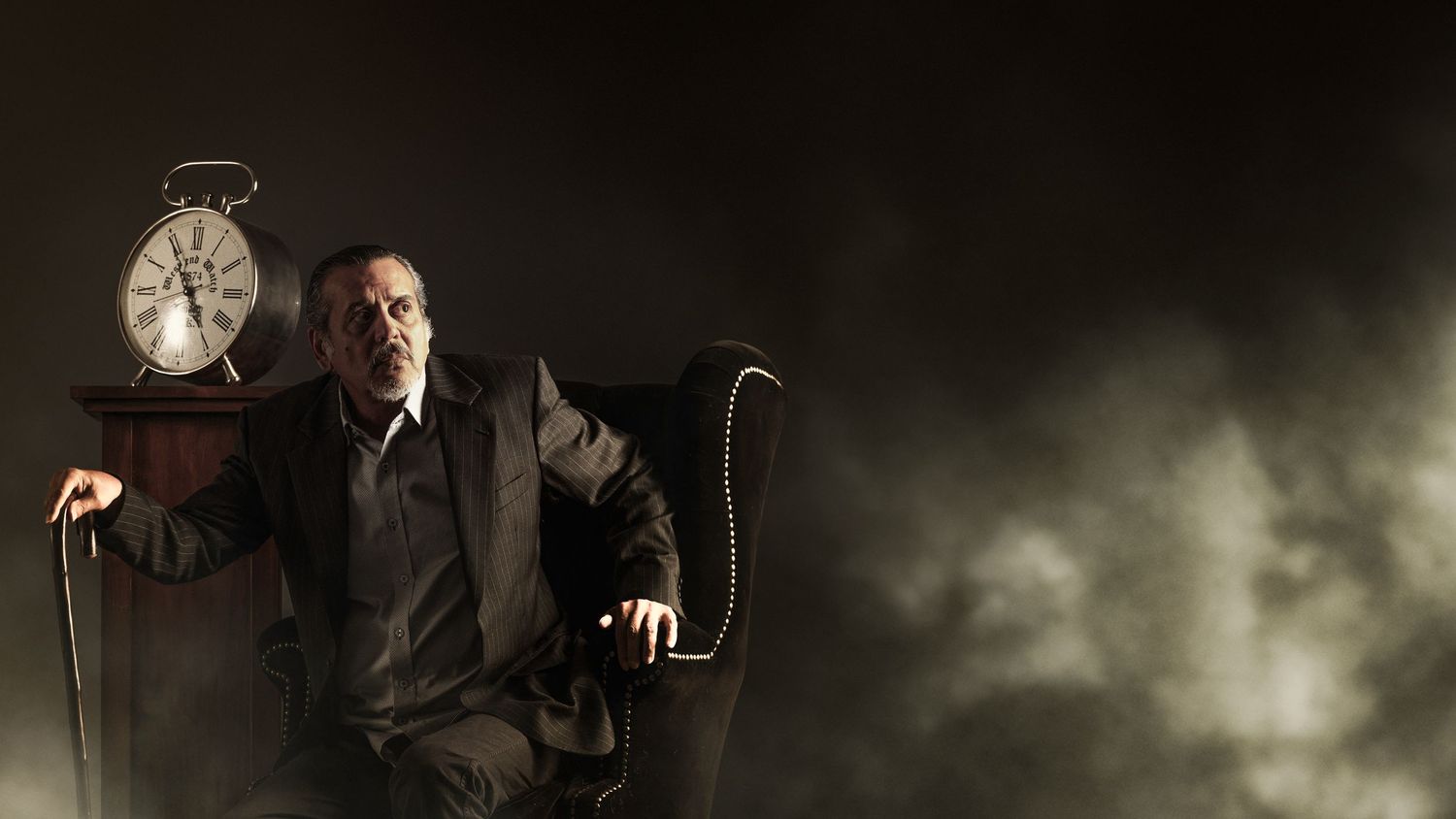Man in dark suit sits in armchair beside vintage clock, dramatic moody lighting with smoke effect.