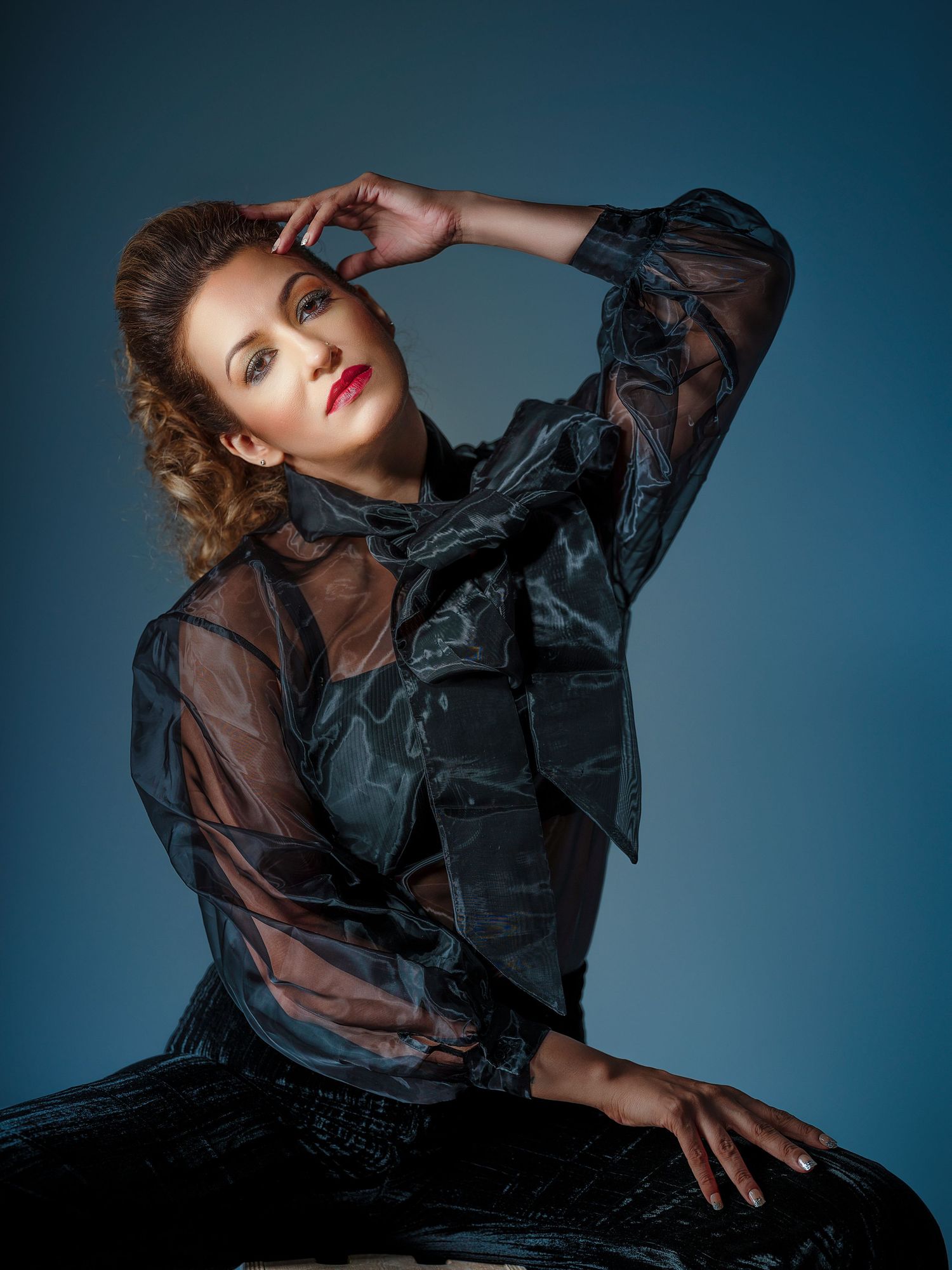 Elegant woman in black sheer blouse posing dramatically against blue studio background.
