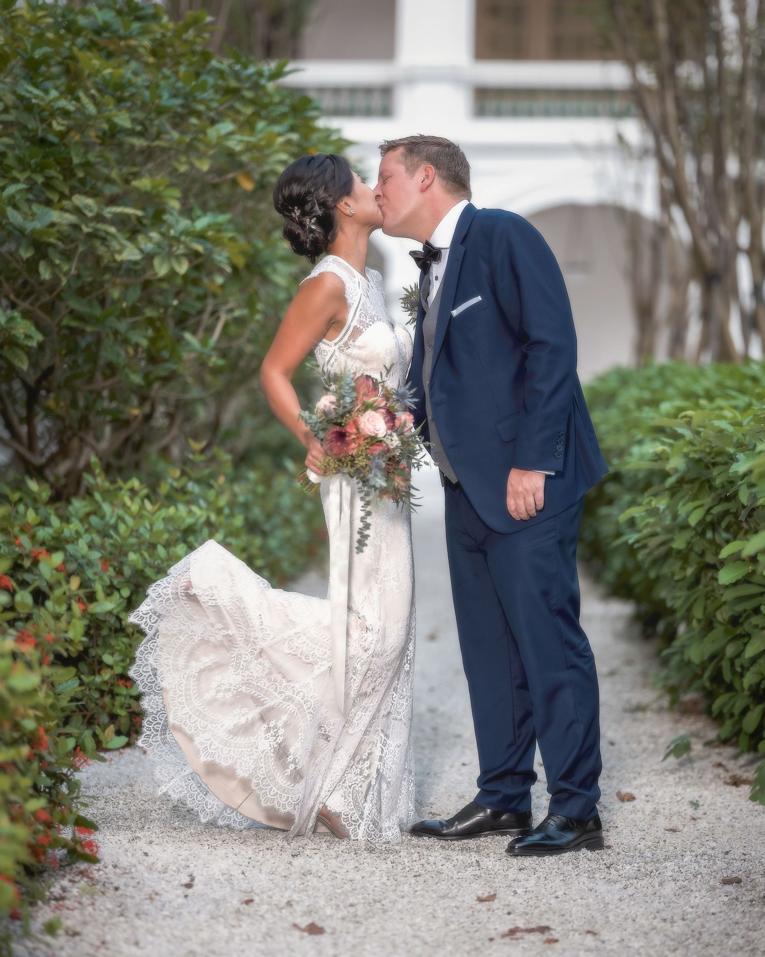 Bride and groom sharing a kiss on garden path, bride in lace gown holding colorful bouquet.