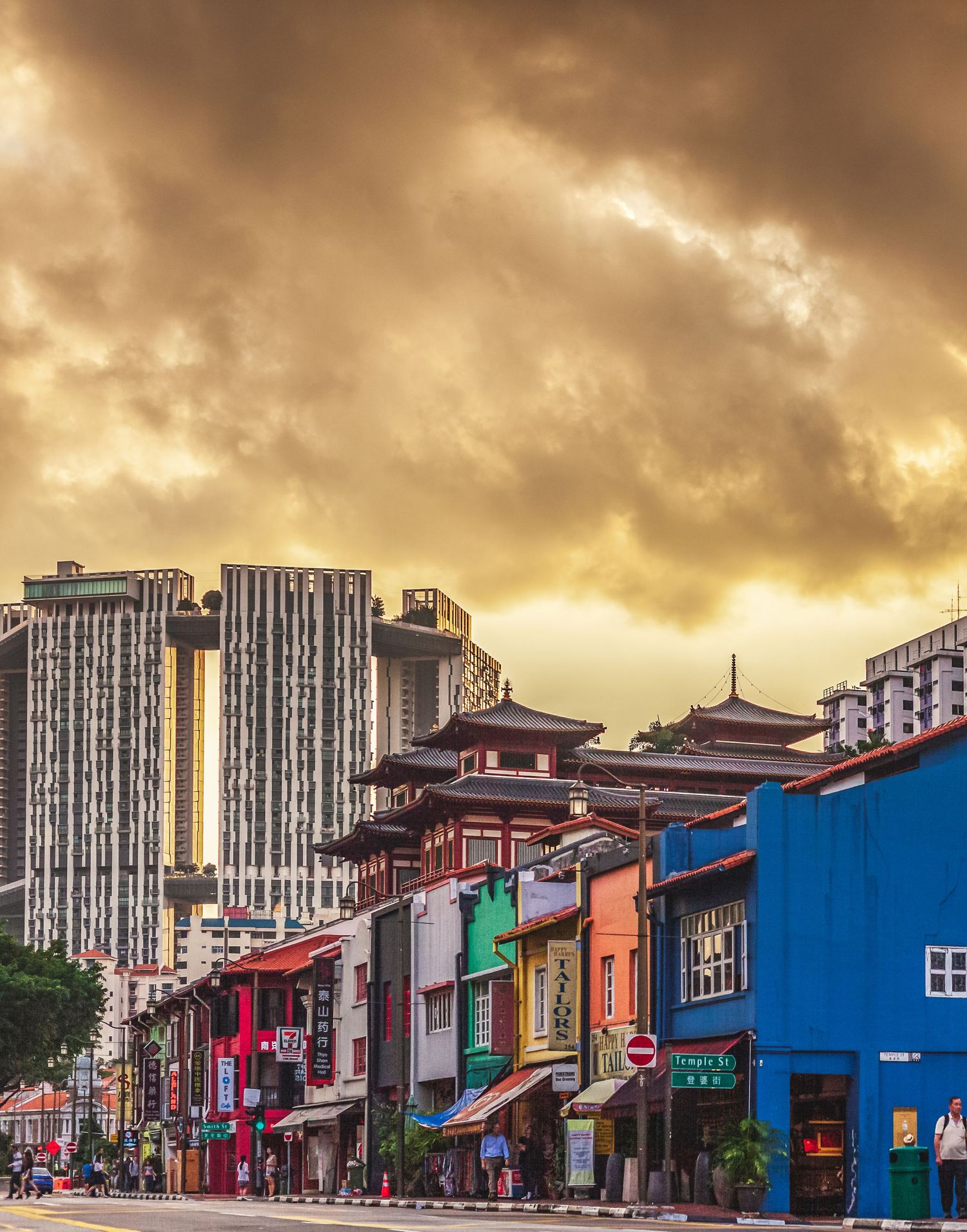 Colorful shophouses line a vibrant street in Singapore's Chinatown district under a dramatic golden sunset sky.