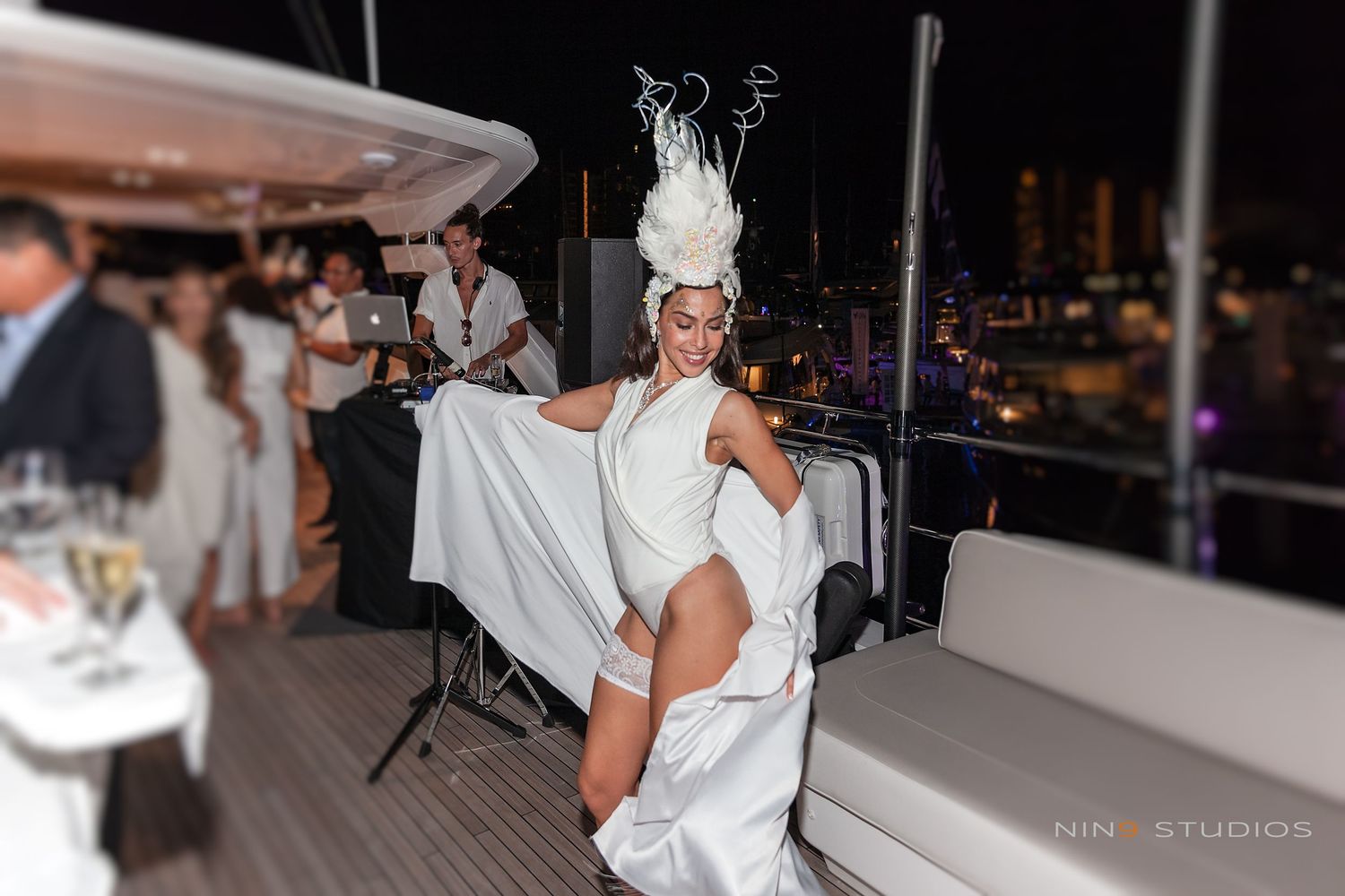 Elegant performer in white dress and feathered headpiece dancing at a rooftop yacht party at night.