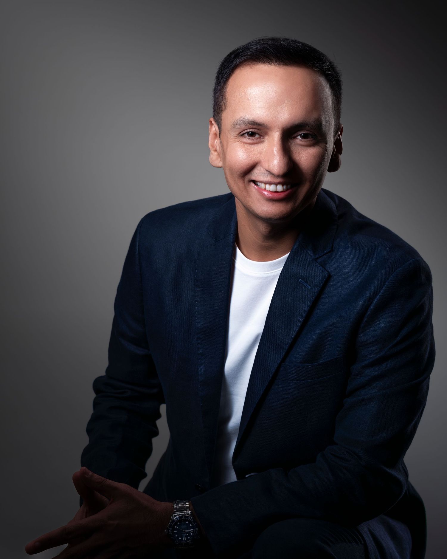 Smiling man in dark blazer and white t-shirt poses confidently with hands clasped in professional headshot photo.