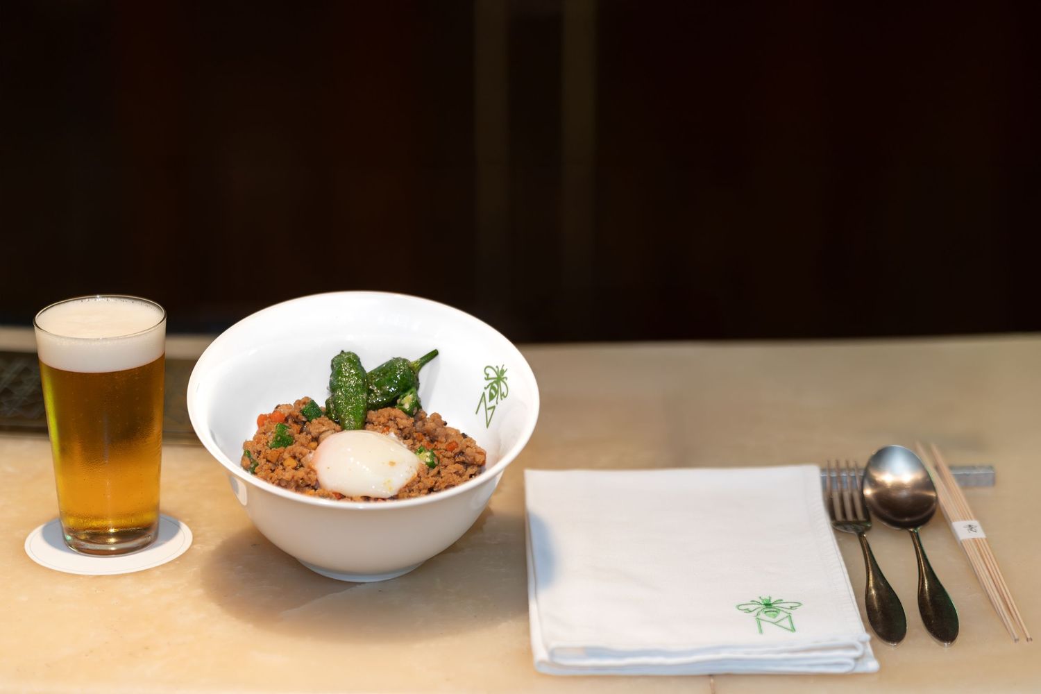 A white bowl of Asian noodles with egg and greens, served with chopsticks, spoon, napkin, and a glass of beer.