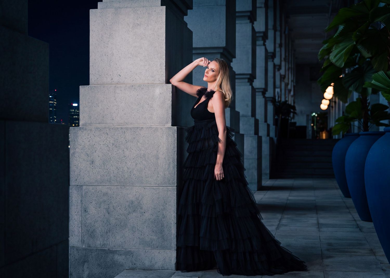 Woman in elegant black tulle gown poses gracefully against stone columns at night.