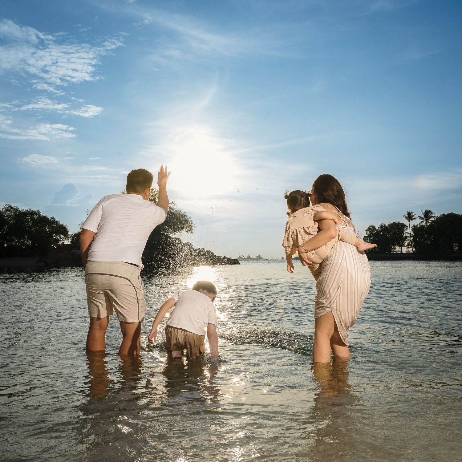 Family of four playing in shallow ocean water on a sunny beach, with parents and two young children splashing joyfully.