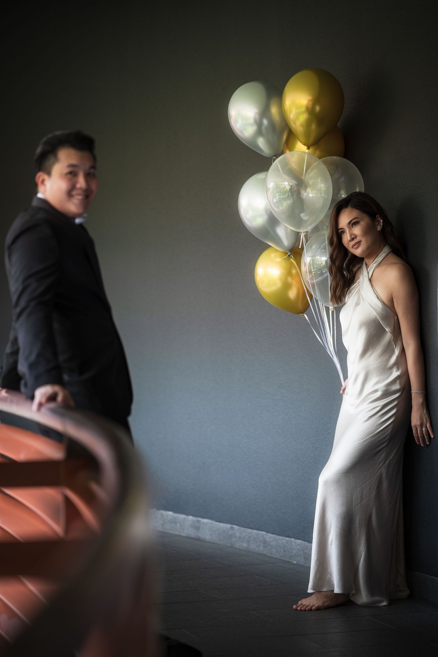 Elegant couple posing with gold and silver balloons, woman in white satin gown leaning against gray wall on staircase.