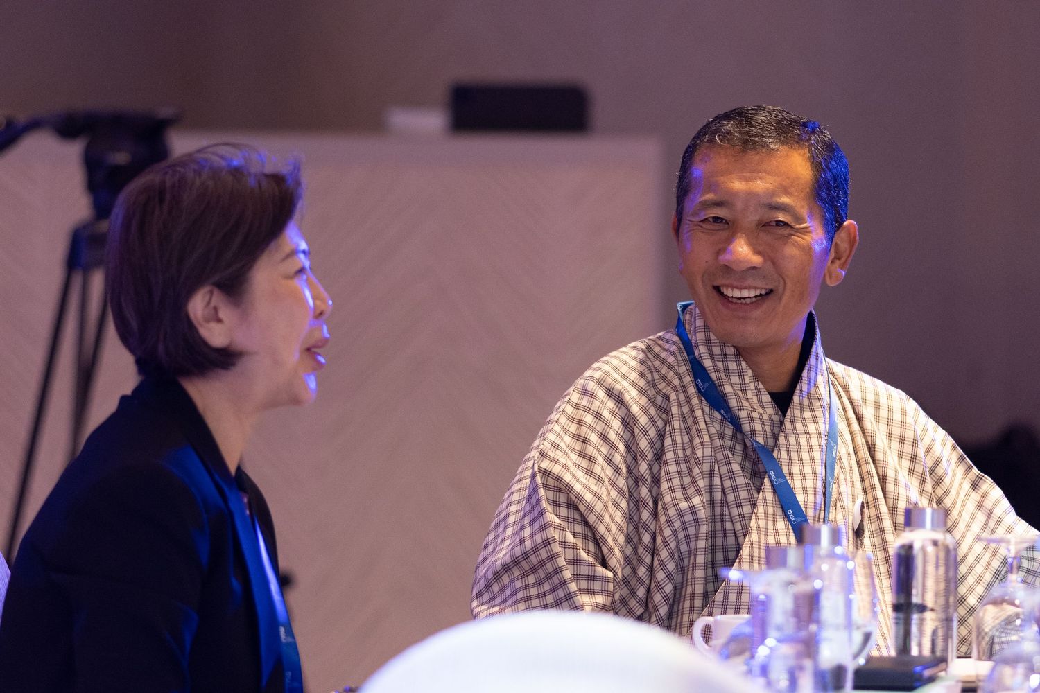 Two professionals smiling and conversing at a conference table with glasses and water bottles.