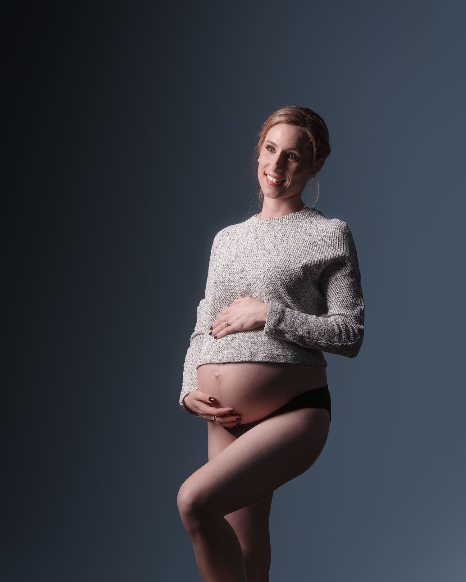Smiling pregnant woman in gray sweater and black underwear cradling baby bump in professional studio portrait.