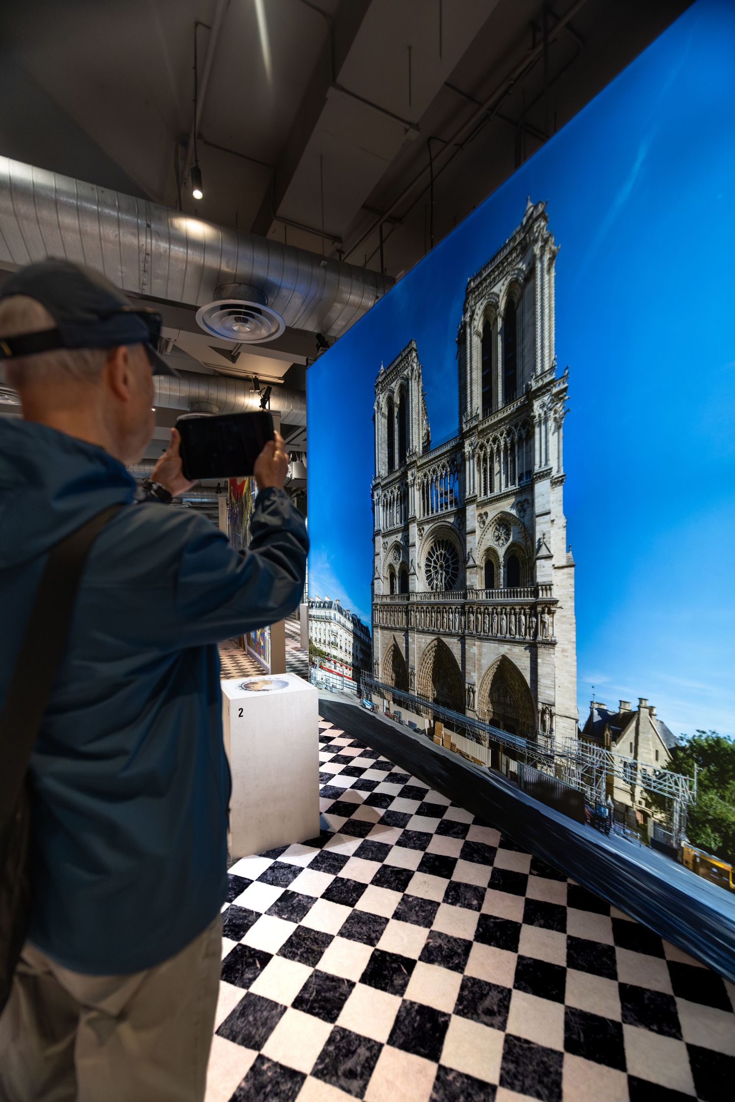 Visitor photographing a large print of Notre Dame Cathedral on display inside an exhibition with checkered flooring.