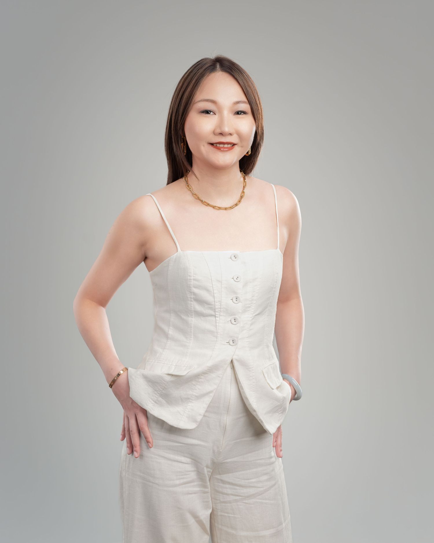 Woman in white linen corset top and pants with gold necklace posing confidently against gray background.