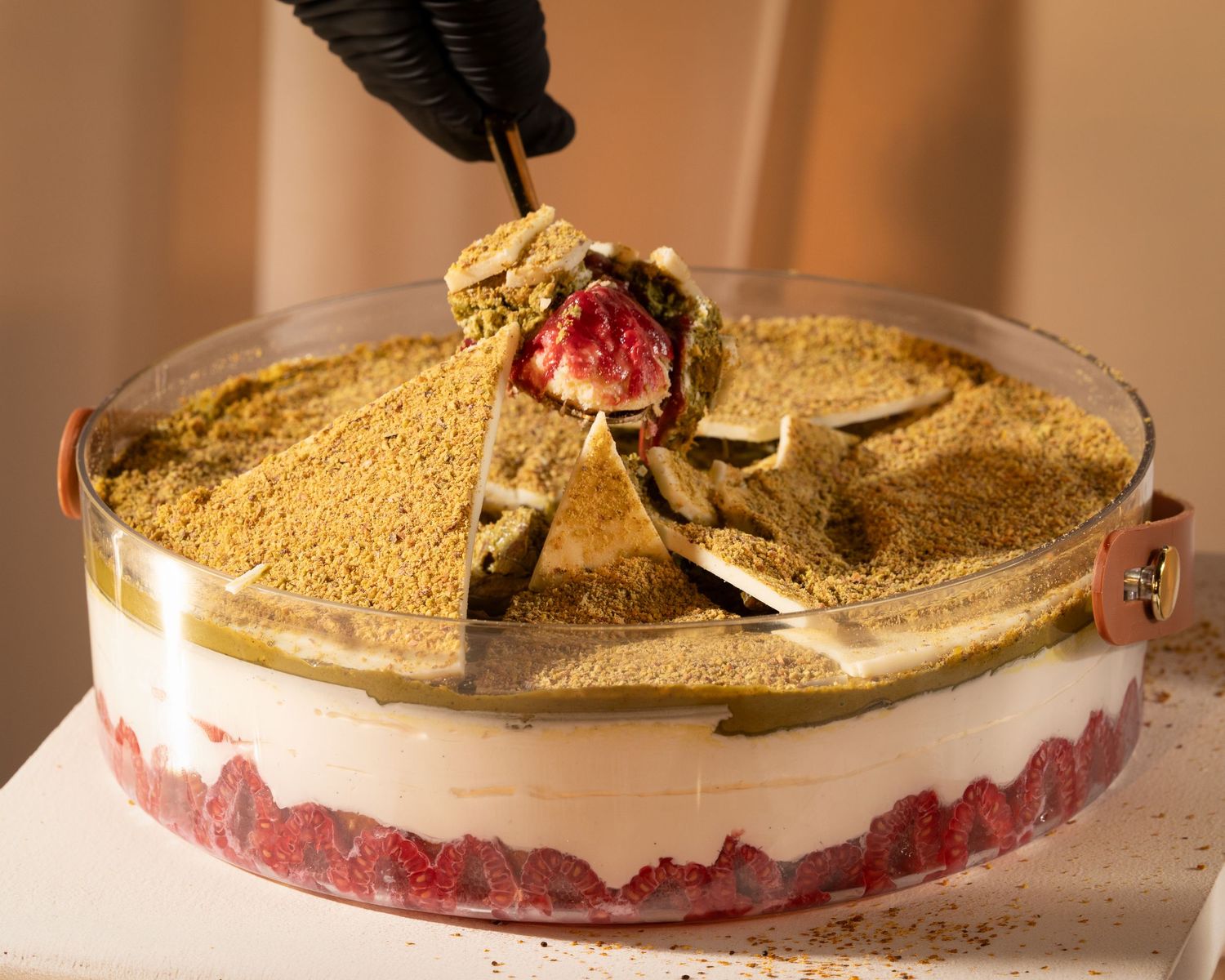 A layered dessert trifle topped with pistachio crumble and raspberries, being served with a spoon in a glass dish.