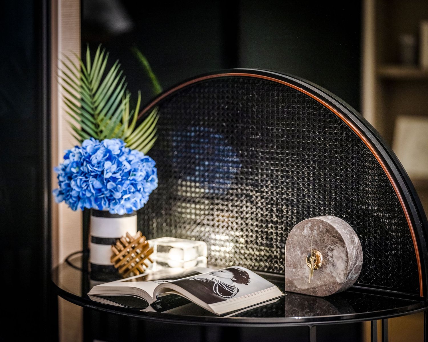 Elegant black side table with blue hydrangea, open book, rattan panel backdrop, and decorative gold accents.