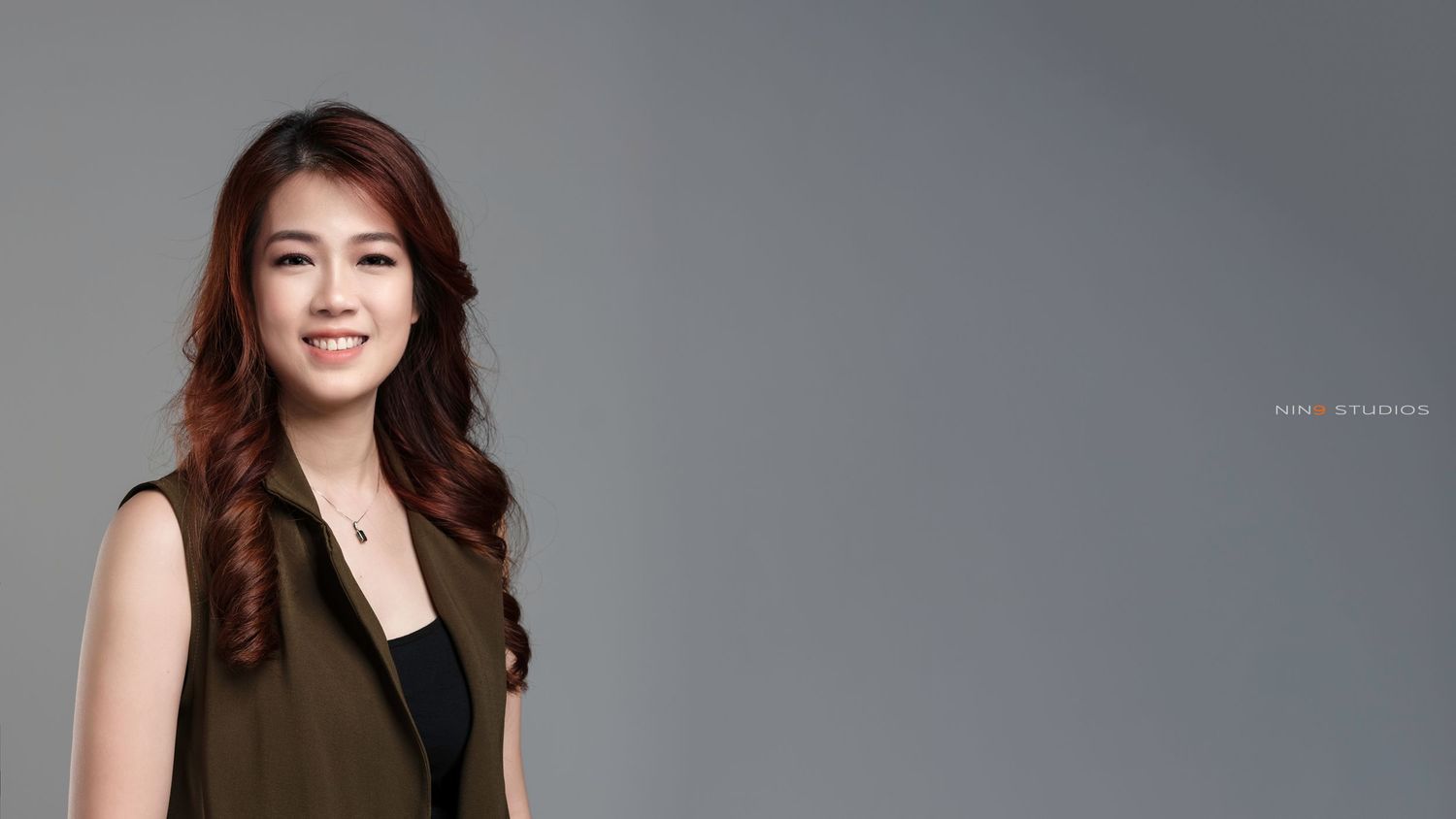 Young Asian woman with long auburn hair smiling confidently against a gray background in a sleeveless olive top.