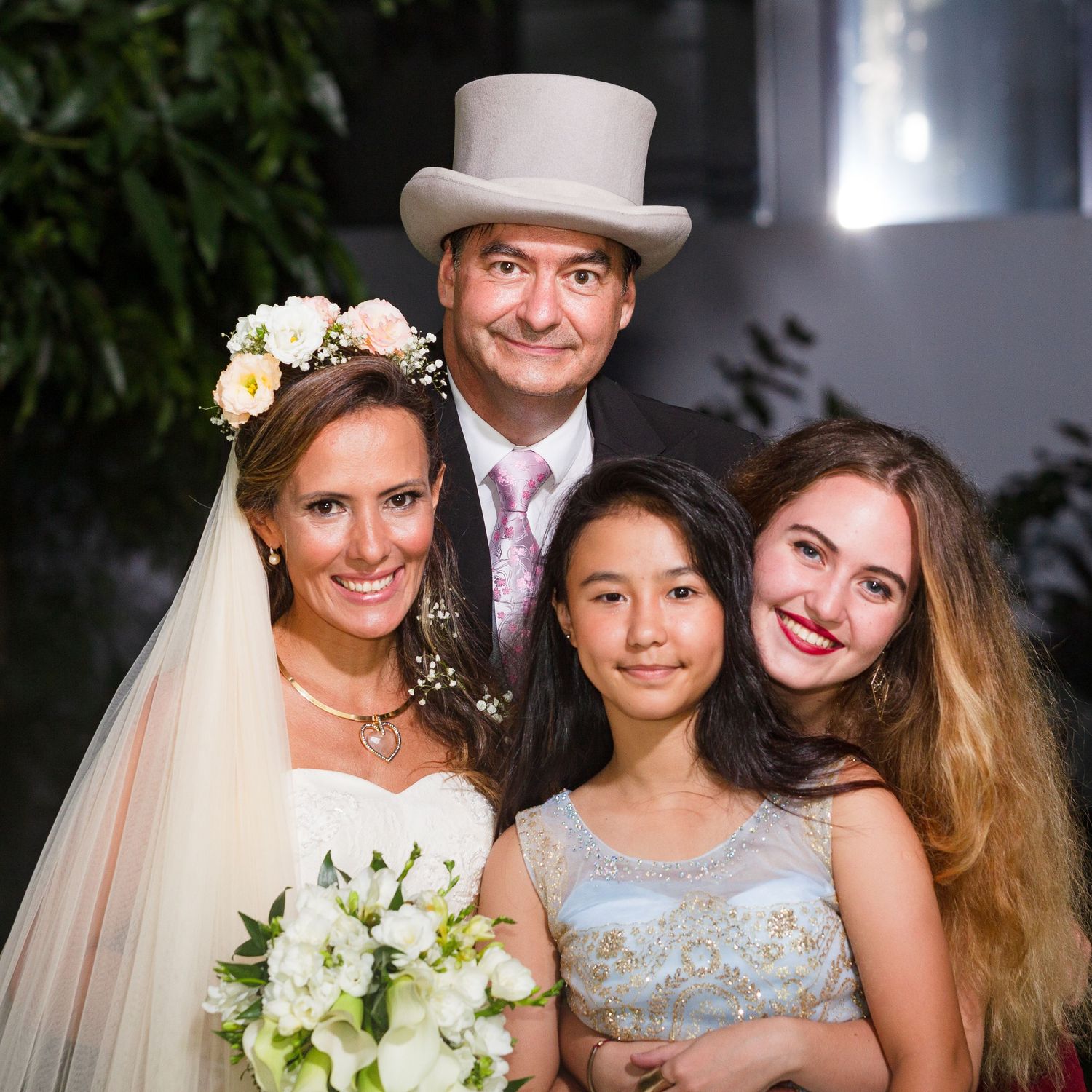 Wedding couple posing with two young girls, bride holding white bouquet, groom in white top hat, formal attire.