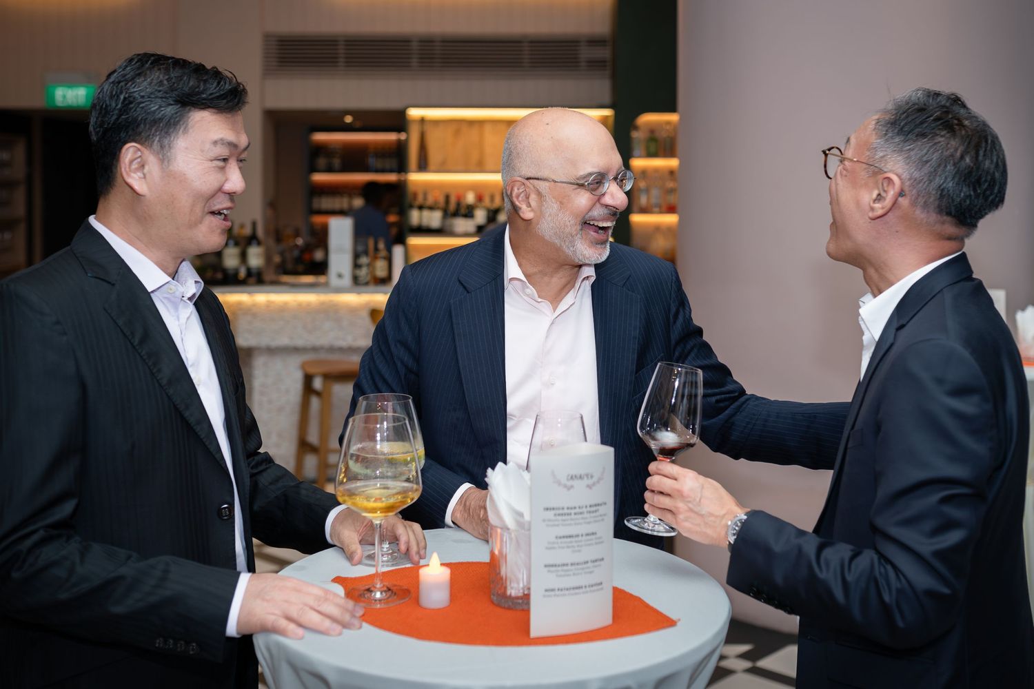 Three men in formal attire smile and toast with wine glasses at an elegant indoor event.