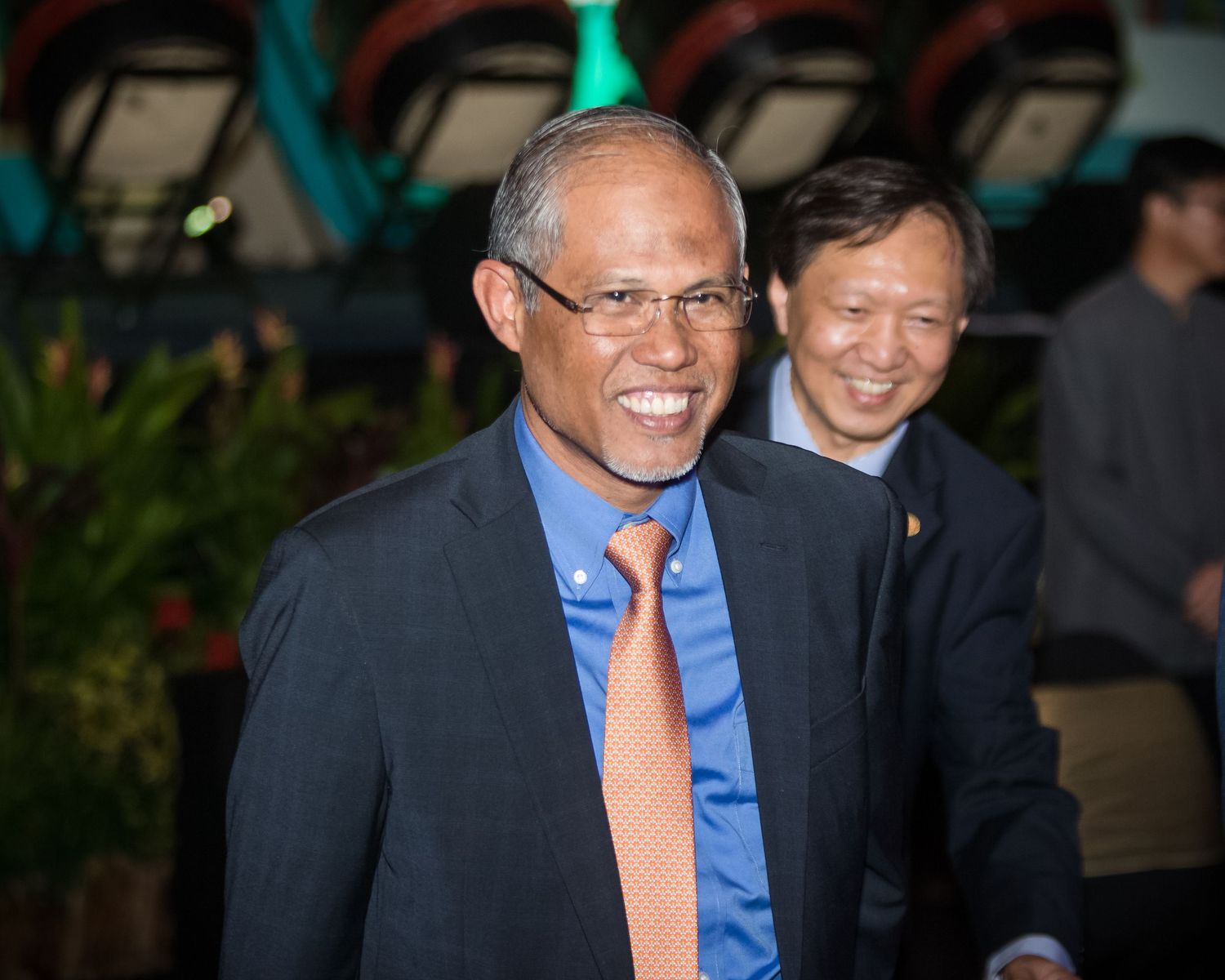 A smiling man in a blue suit and orange tie walks at a formal event with another man behind him.