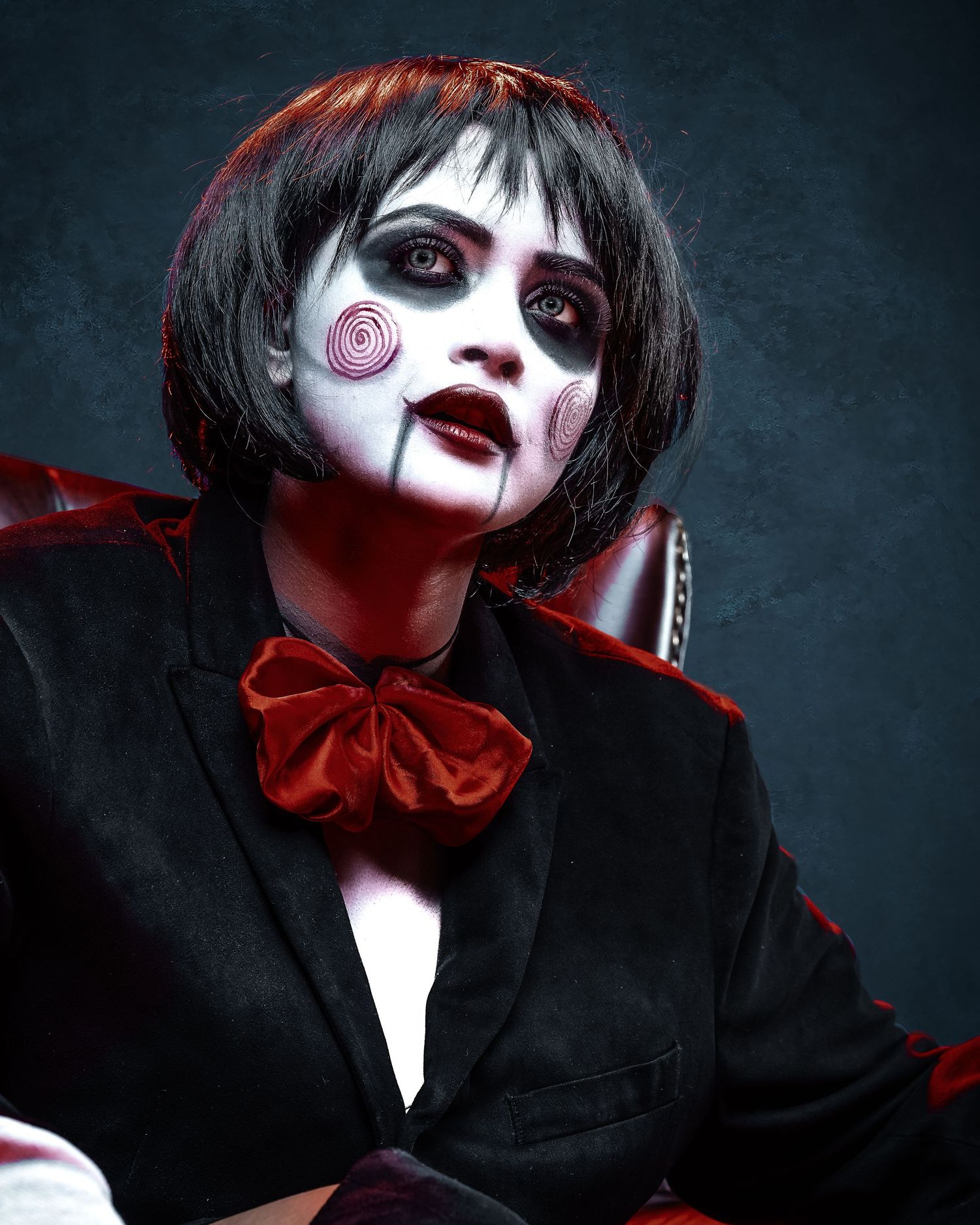 Person in Billy the Puppet cosplay with white face paint, red spiral cheeks, black bob wig, suit, and red bow tie.