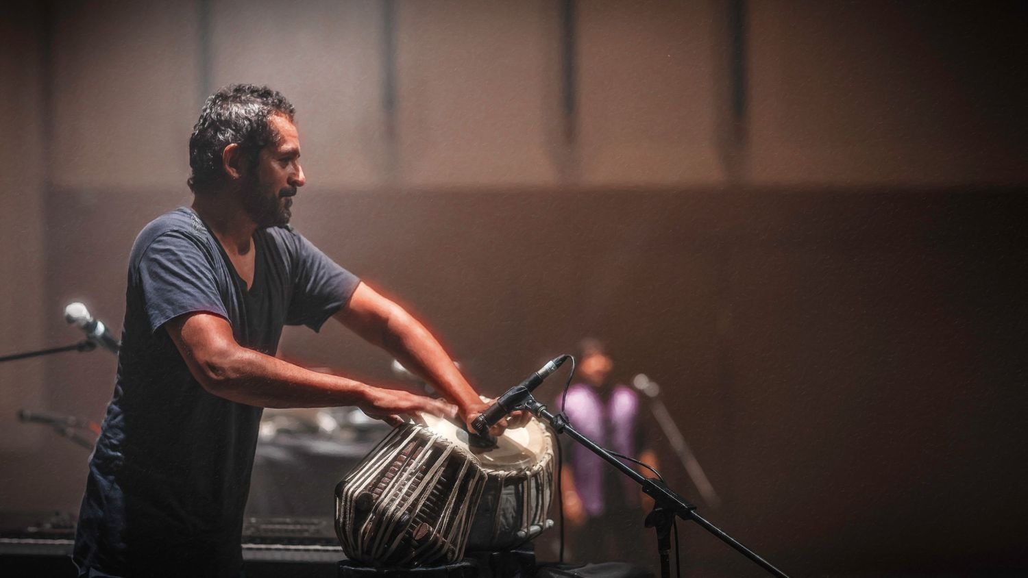 Musician playing tabla drums on stage with microphones in a dimly lit performance venue.