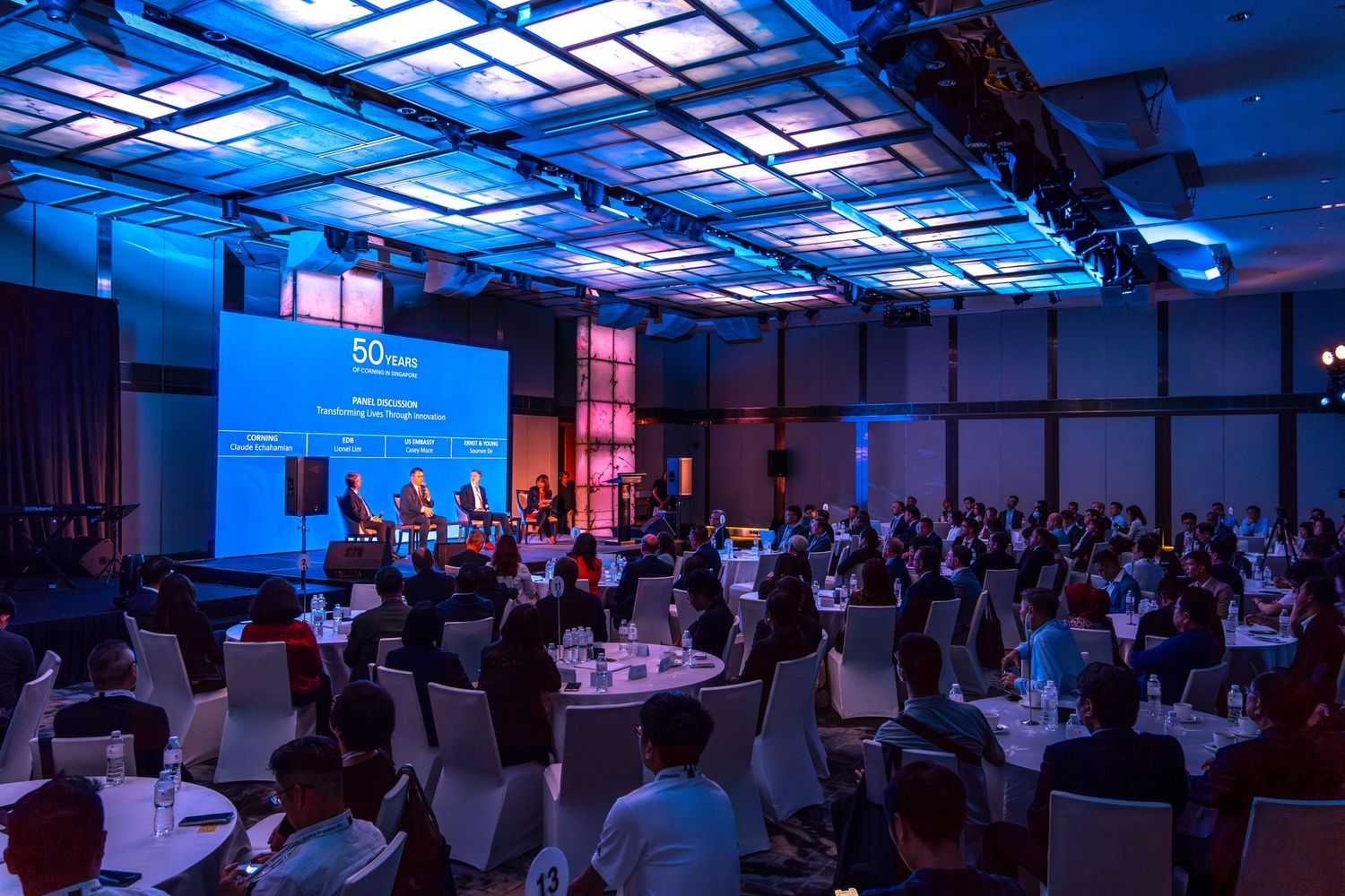 Large corporate conference hall with audience seated at round tables under blue lighting.