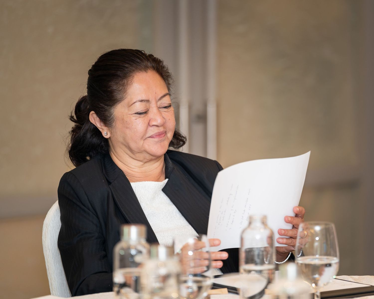 A woman in a black blazer reads documents at a conference table with glasses and water bottles in the foreground.