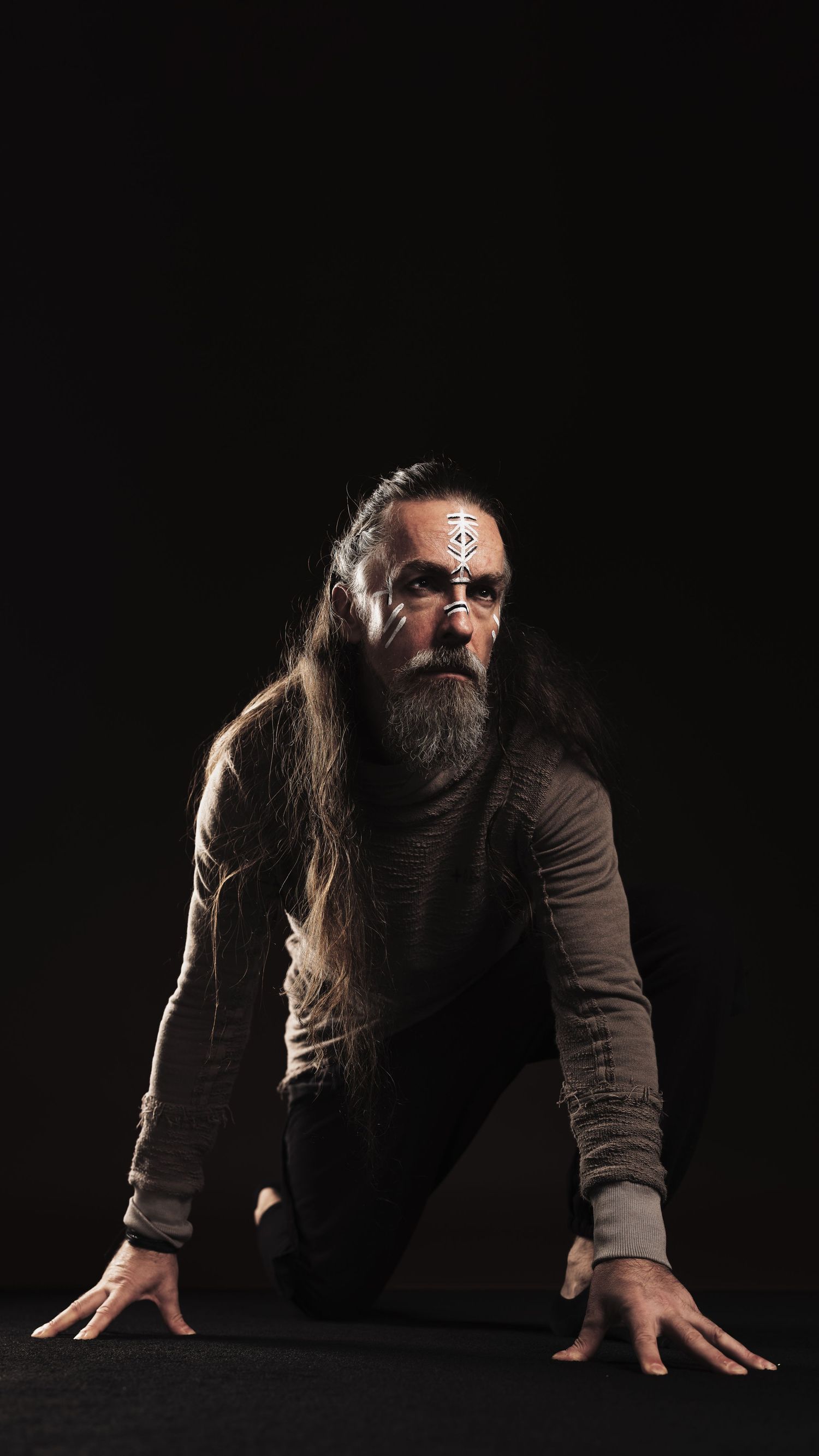 Long-haired bearded man with face paint crouches on black floor in dramatic dark studio portrait.