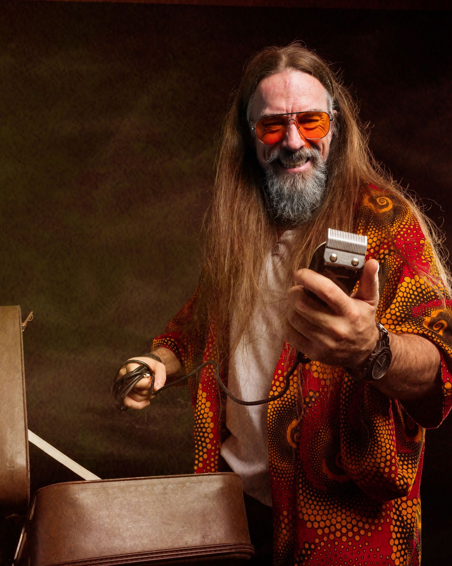 Long-haired bearded man in colorful orange patterned robe and tinted glasses holding a small device, smiling warmly.