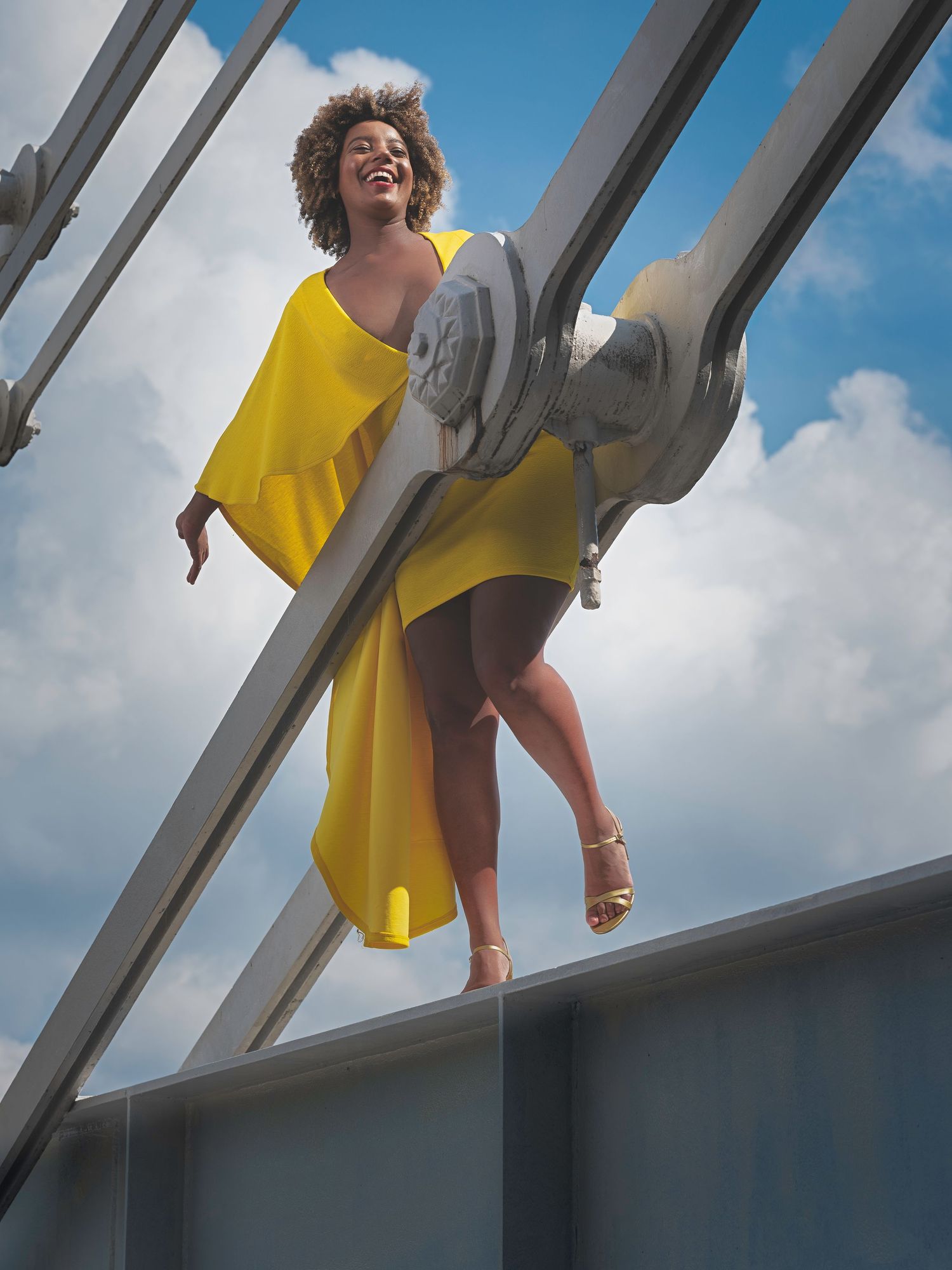 Joyful woman in yellow dress poses on a modern bridge structure against a blue sky backdrop.