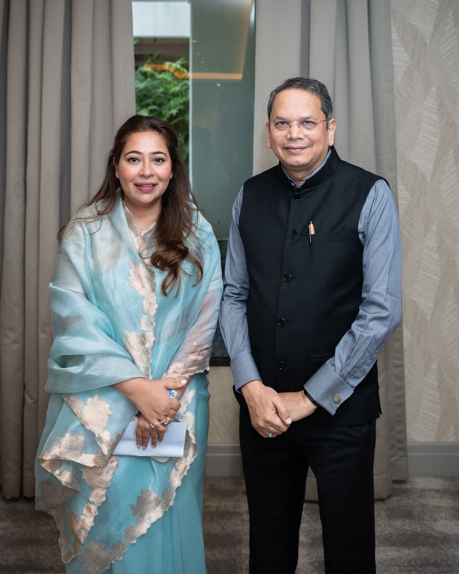 A woman in a light blue traditional outfit and a man in a black vest pose together for a formal photo indoors.