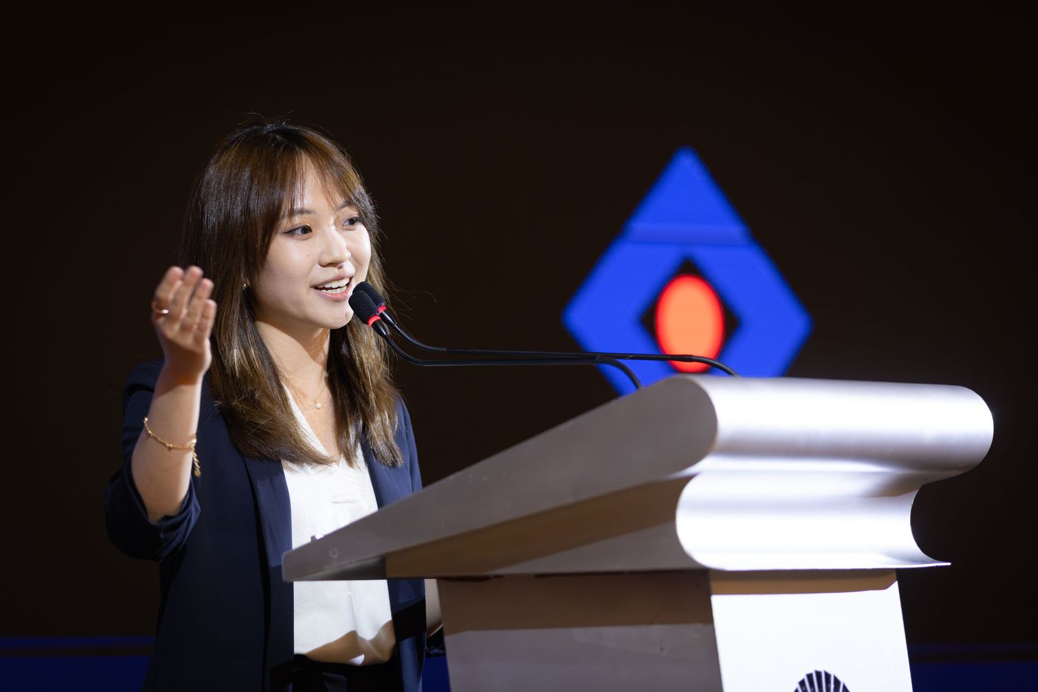 Young Asian woman in blazer delivering speech at podium during formal presentation or conference.