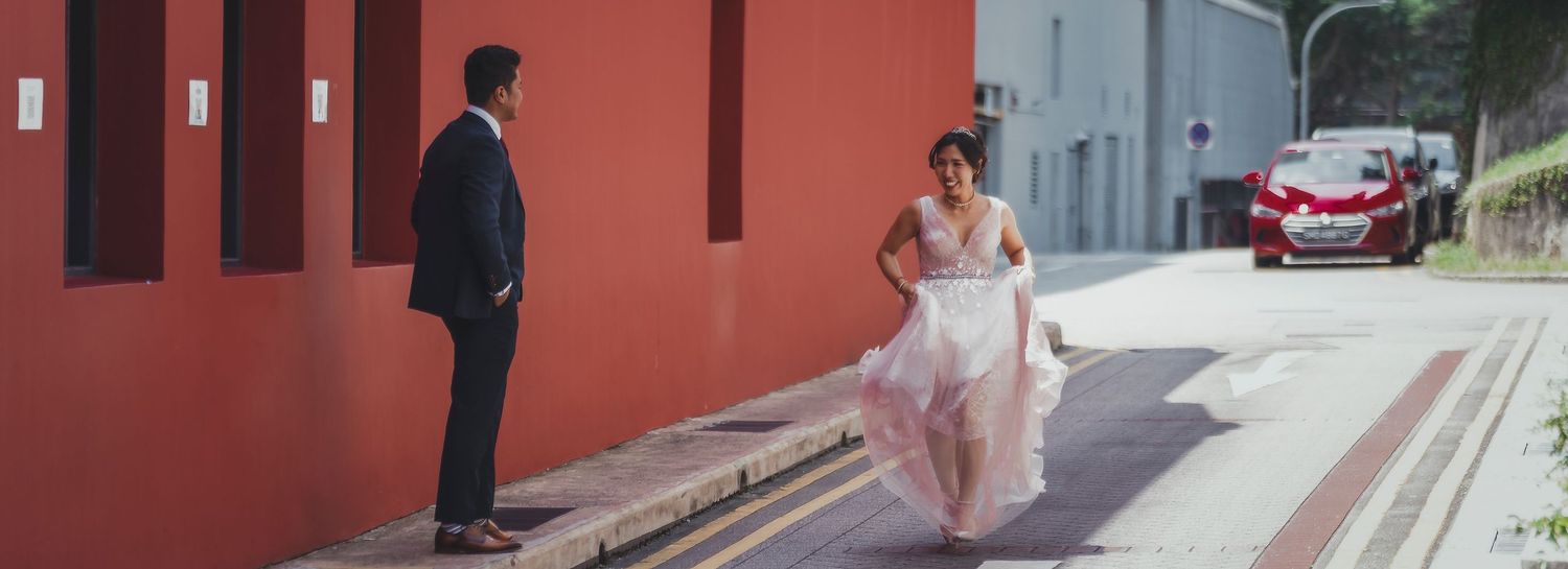 A bride in a flowing white gown walks toward a groom in a black suit on a street beside a vibrant red wall.