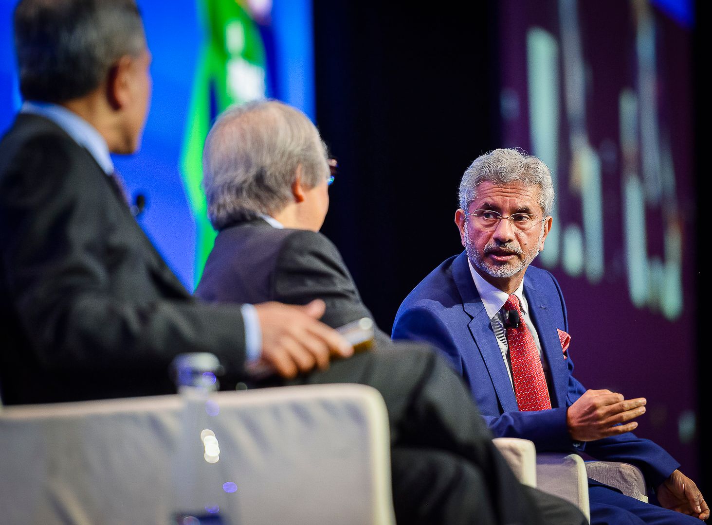 Three men in suits engage in a panel discussion on stage at an international summit conference.