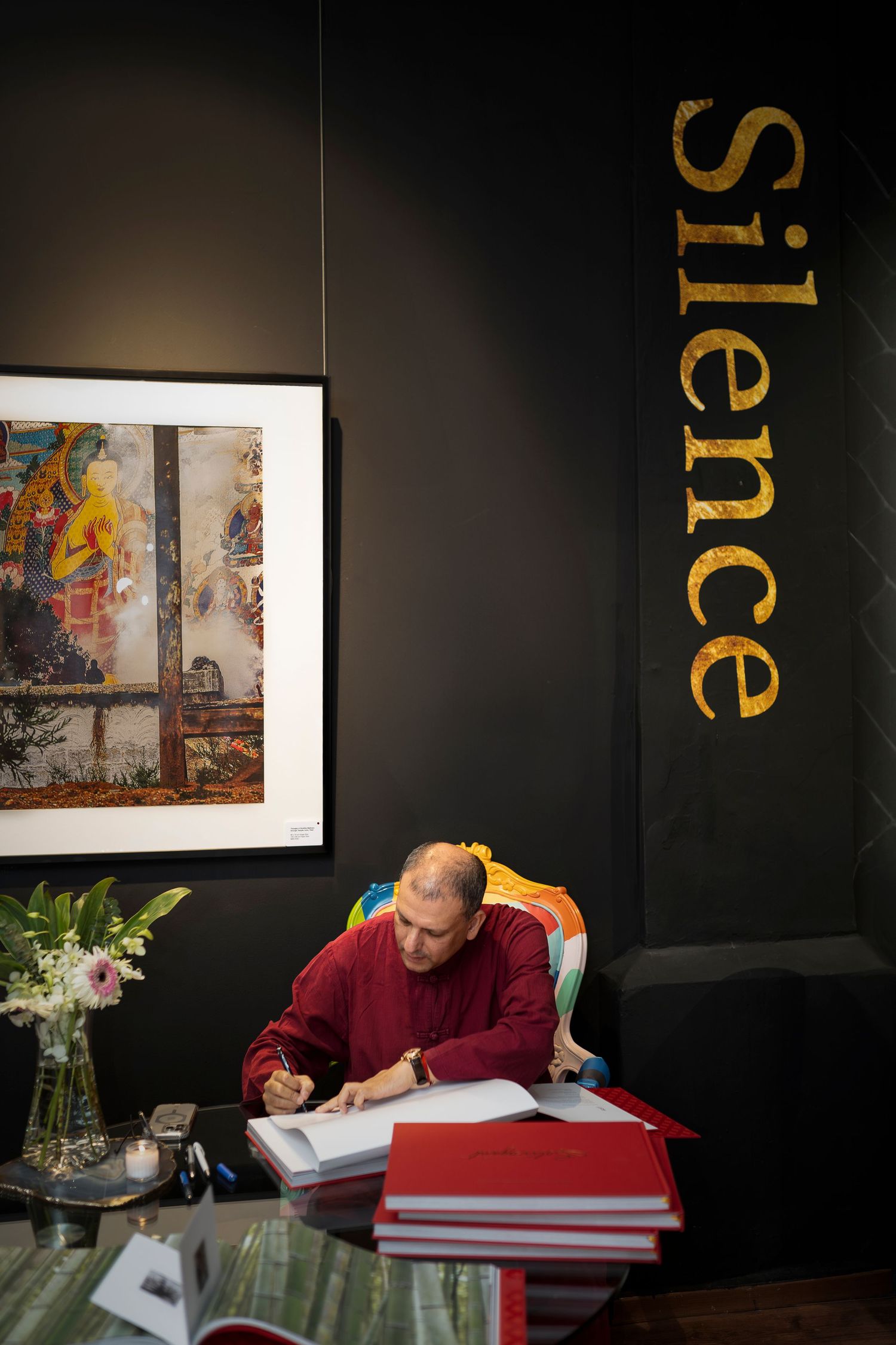 Monk in red robes writing at a desk beneath a gold Silence sign in a dark gallery space.