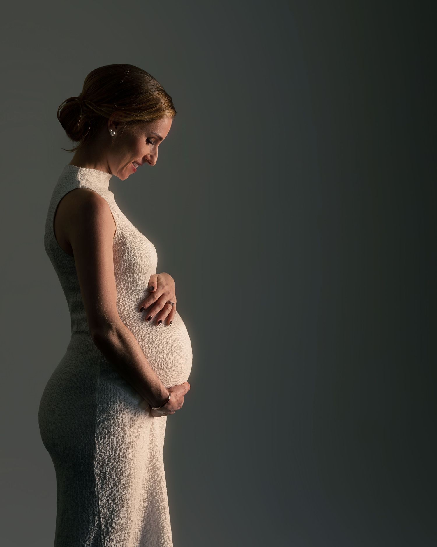 Elegant pregnant woman in white dress cradling baby bump, side profile maternity portrait on gray background.