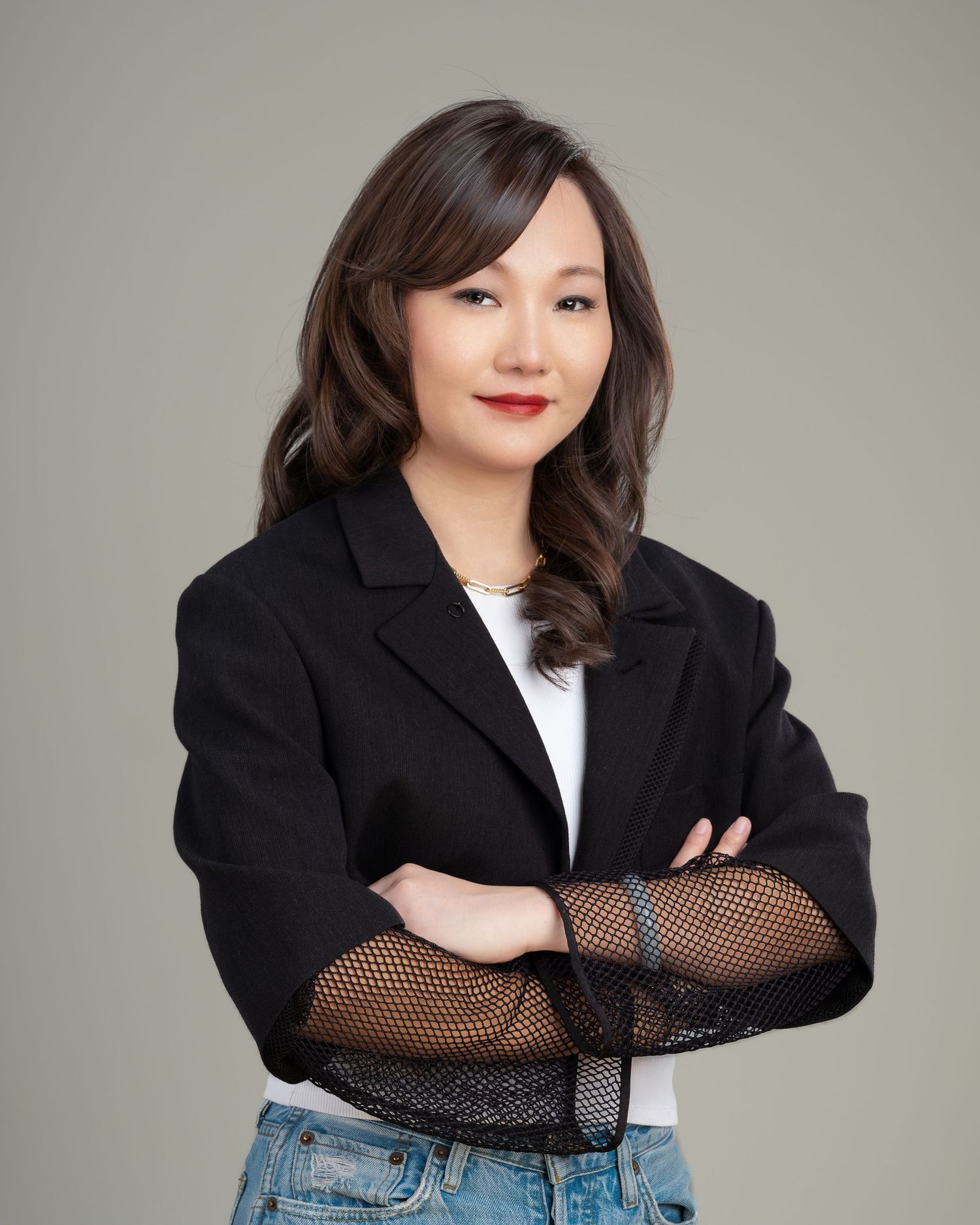 Asian woman in black blazer with arms crossed posing confidently against neutral studio background.