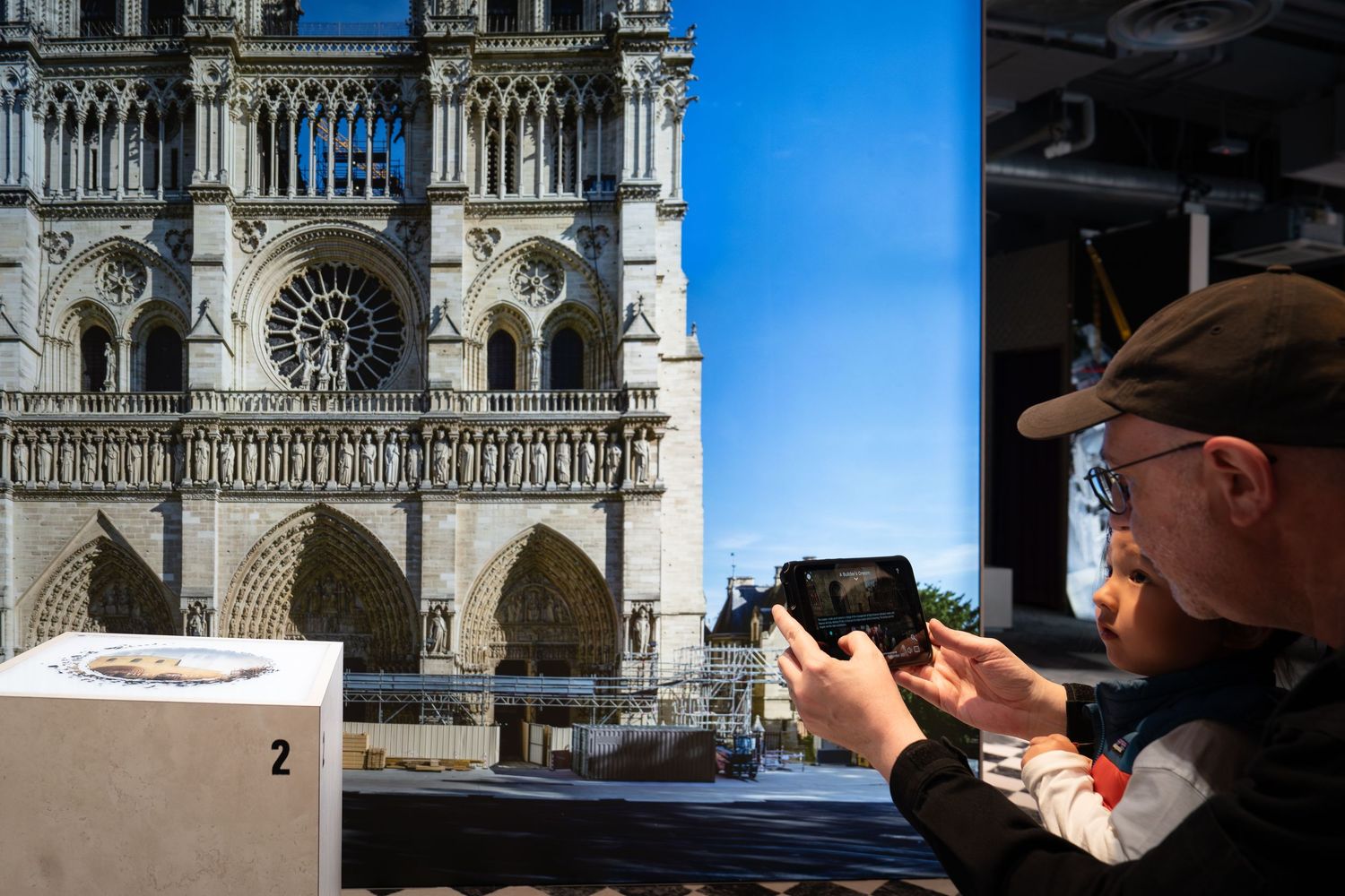 Man photographs Notre Dame Cathedral facade displayed on a large screen at an immersive exhibition.