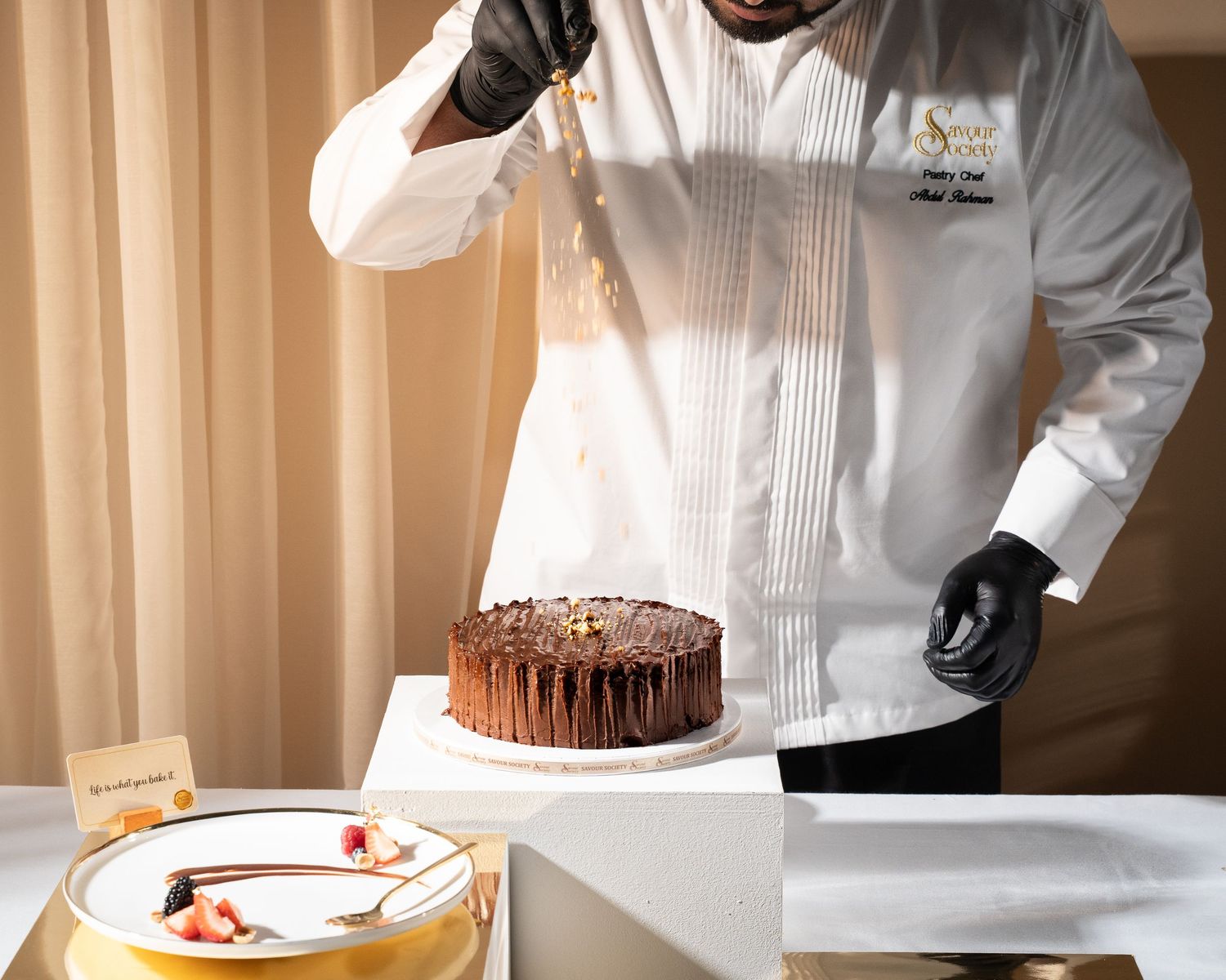 A pastry chef in white uniform decorating a chocolate layer cake with gold drizzle on a white stand.