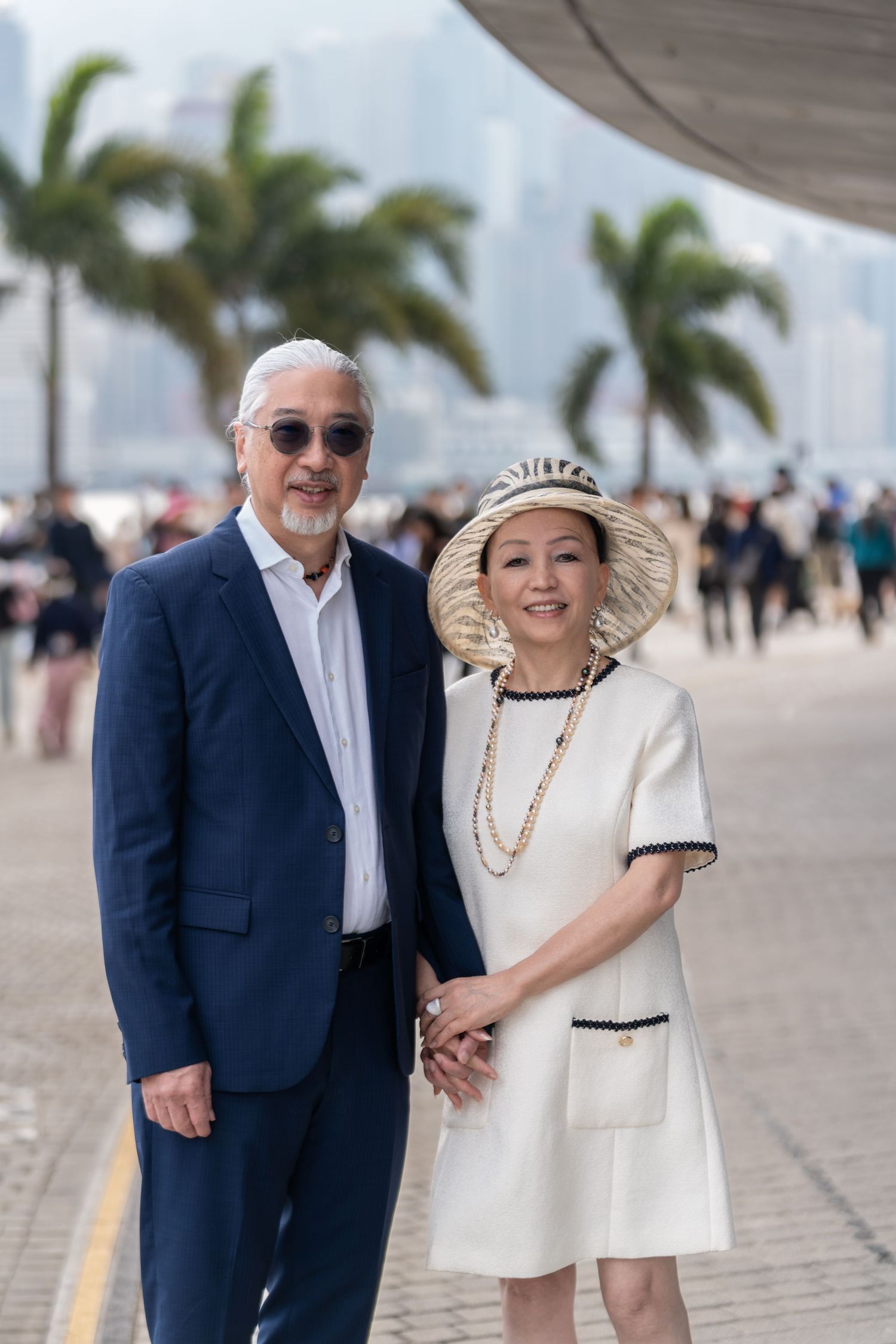 Elegant senior Asian couple holding hands on a waterfront promenade with palm trees and city skyline.