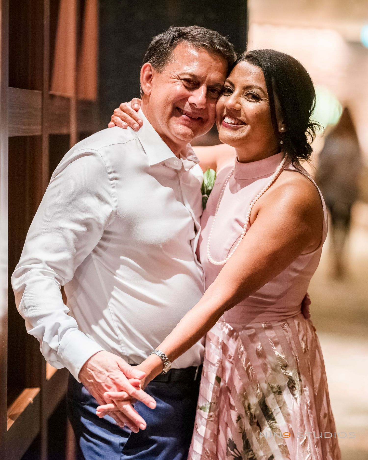 A smiling couple poses warmly together, woman in pink floral dress embracing man in white shirt at an elegant event.