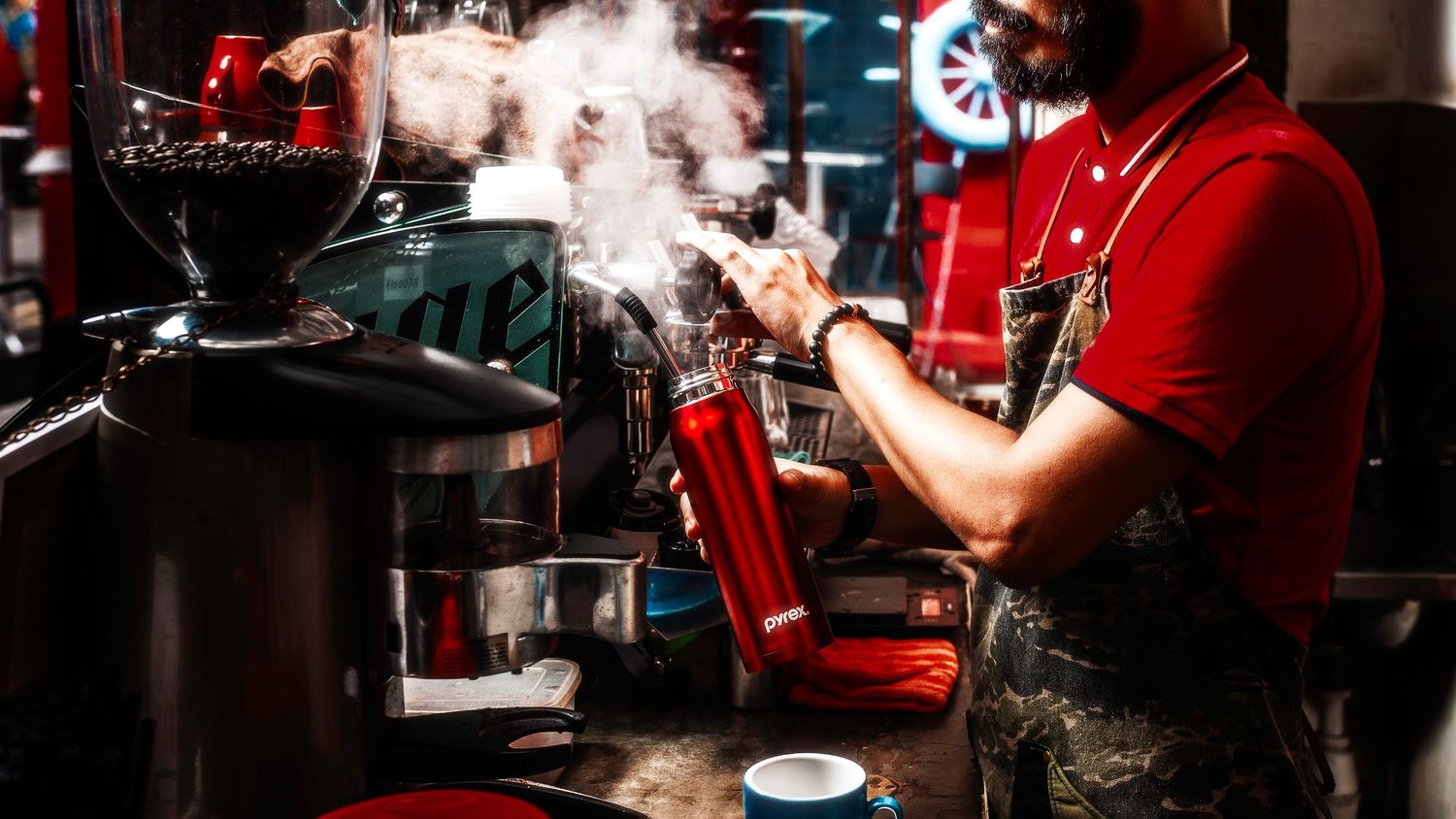 Barista in red uniform operating an espresso machine with steam rising in a moody coffee shop setting.