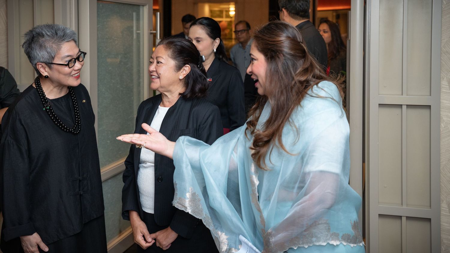 Three women in elegant attire share a joyful moment, one wearing a light blue shawl, at a formal indoor gathering.