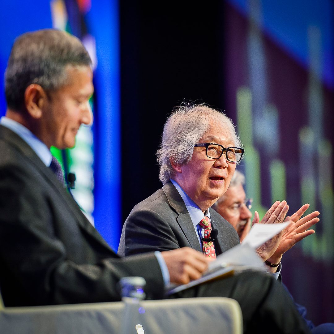 Three panelists engage in discussion at a formal business conference, with the central speaker gesturing expressively.