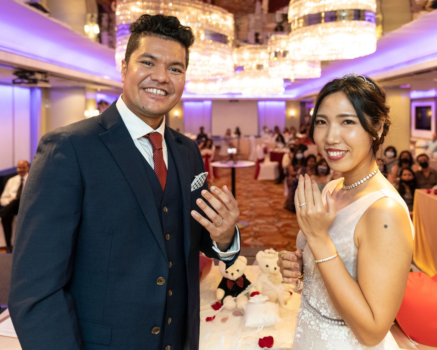 Couple in formal attire smiling and showing rings at elegant wedding reception venue with chandeliers.