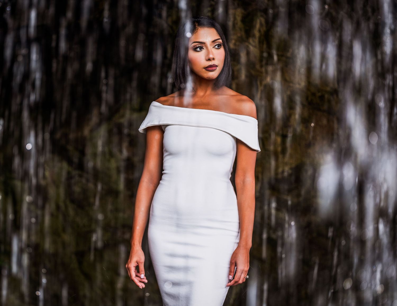 Woman in white off-shoulder bodycon dress posing elegantly in front of a blurred waterfall backdrop.