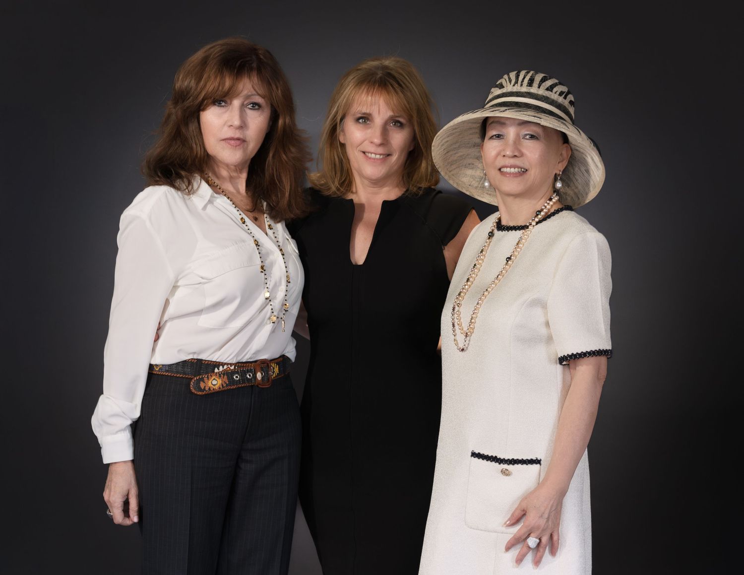 Three elegantly dressed women pose together, one wearing a stylish hat, in a professional portrait photo.
